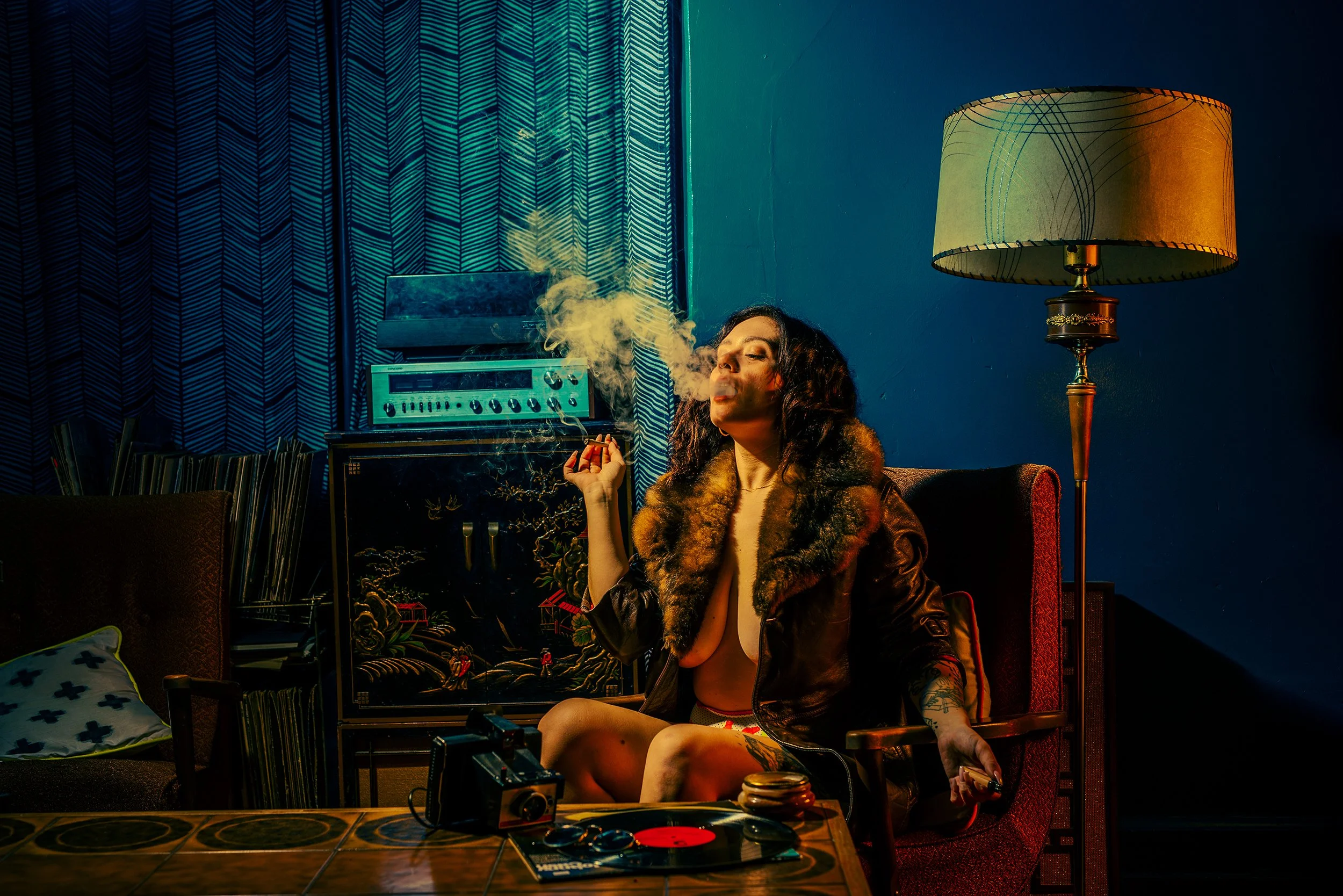 A woman with dark curly hair sitting on a red velvet chair in a dimly lit room, exhaling smoke. She is wearing a leather jacket with a fur collar, and has tattoos on her arms. The room has vintage decor, a table with vinyl records, an old radio, a ca