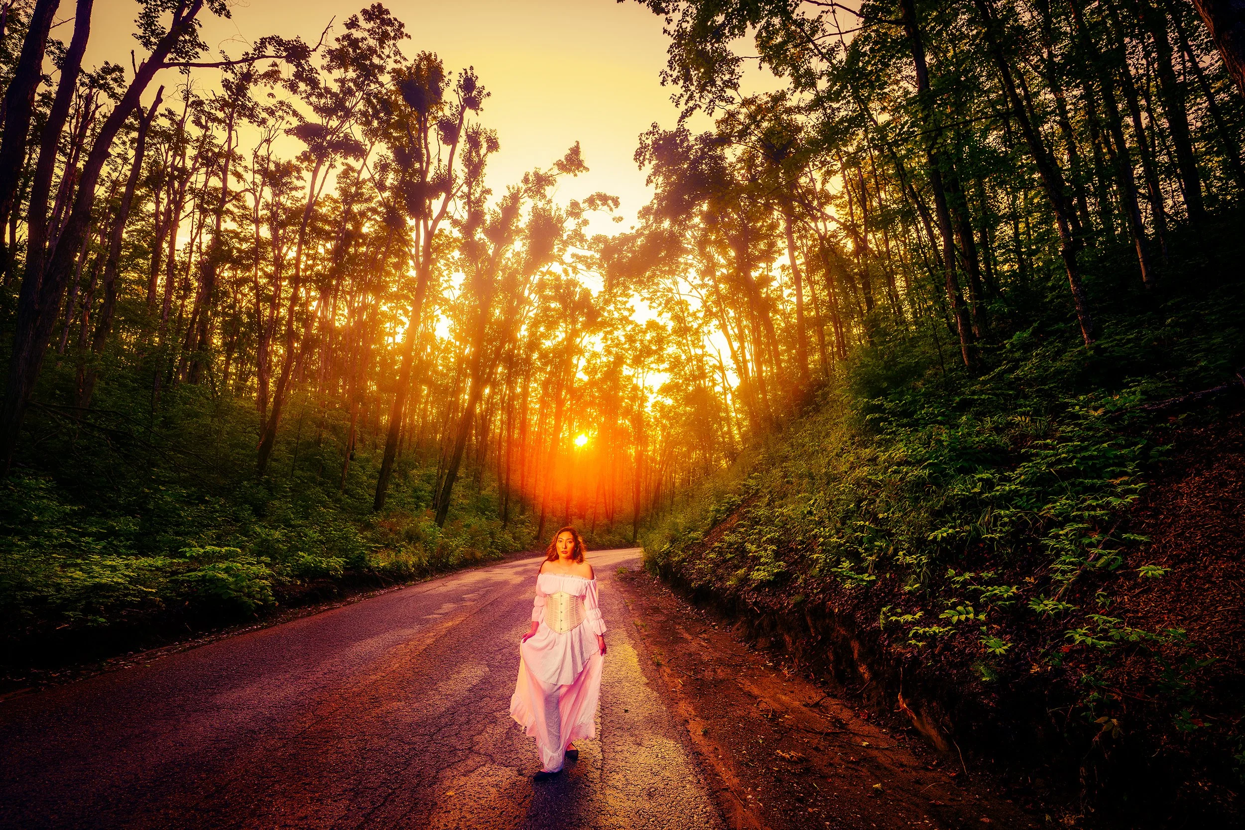A woman in a white dress walking on a deserted road surrounded by a dense forest during sunset.