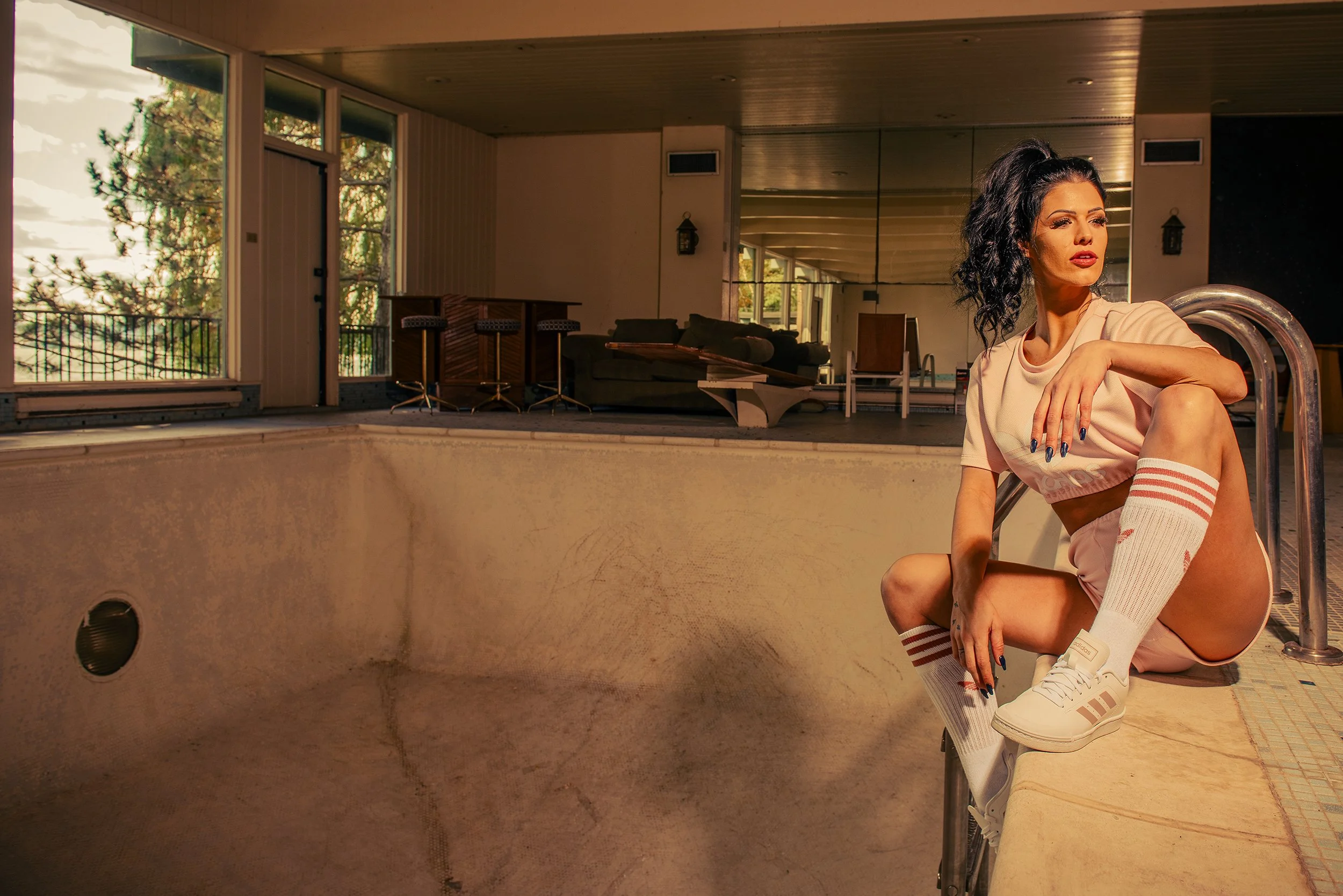 A young woman with dark curly hair sitting by an indoor swimming pool railing, wearing a beige t-shirt, pink shorts, white sneakers, and striped socks. In the background, large windows reveal outdoor trees and sky, with a living space that includes s