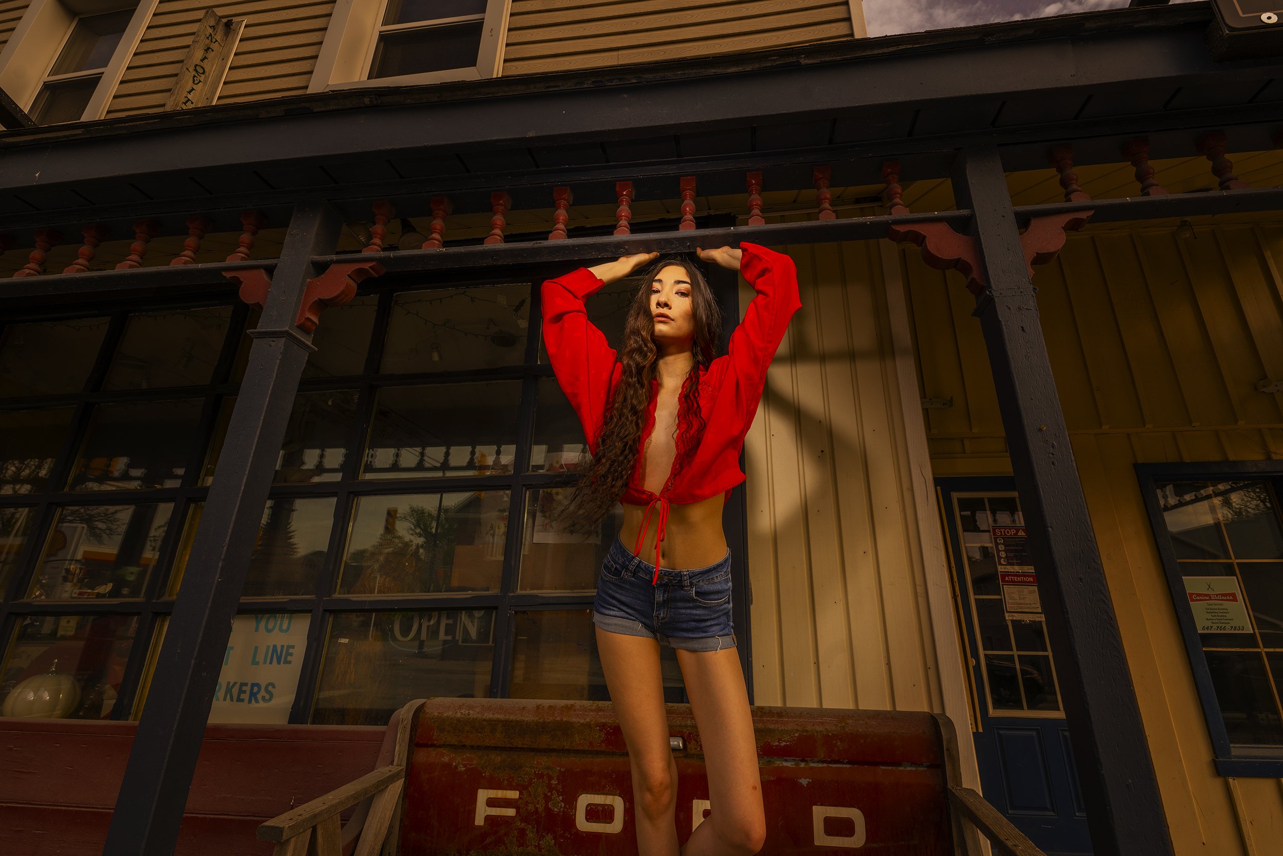 Young woman with long wavy hair and wearing a red top and denim shorts standing on a porch, holding onto a balcony railing, in front of a building with large windows and a sign that says 'OPEN'.