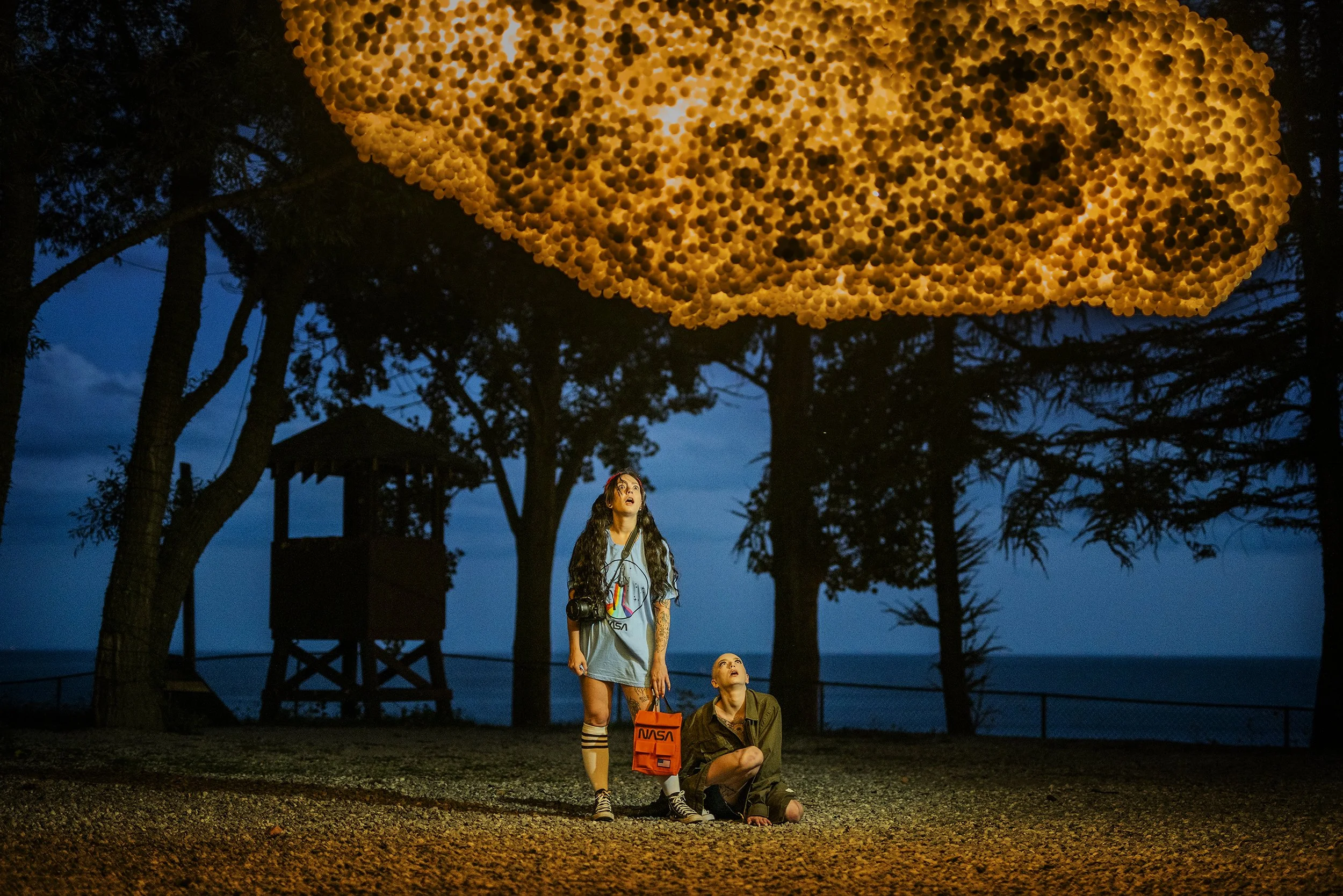Two young women looking up at a large cluster of illuminated lanterns hanging overhead outdoors at dusk, with trees and a lifeguard tower in the background.