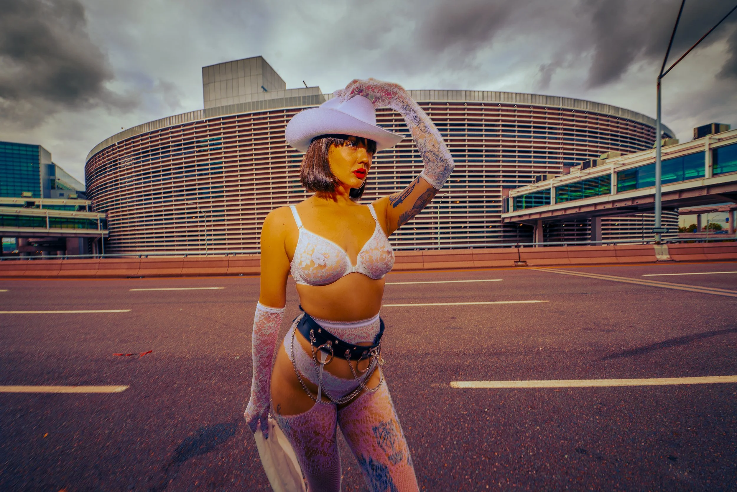 A woman with short dark hair, wearing a white lace bra, sheer lace pants, long lace gloves, a white wide-brimmed hat, and a black belt with chains, standing on an empty city street with a modern curved building and cloudy sky in the background.