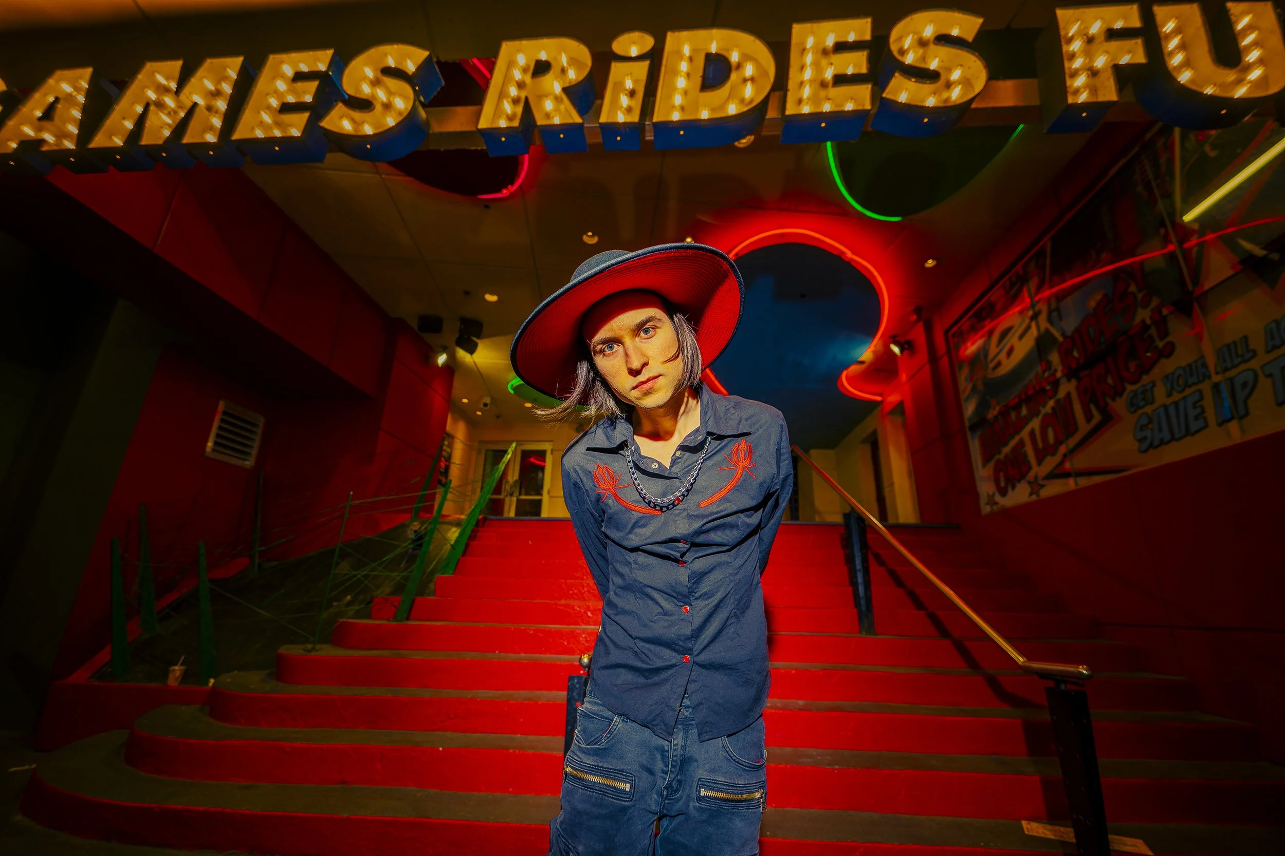 Person with shoulder-length hair wearing a wide-brimmed hat and a button-up shirt standing on red-carpeted stairs inside a venue with neon and illuminated signs.