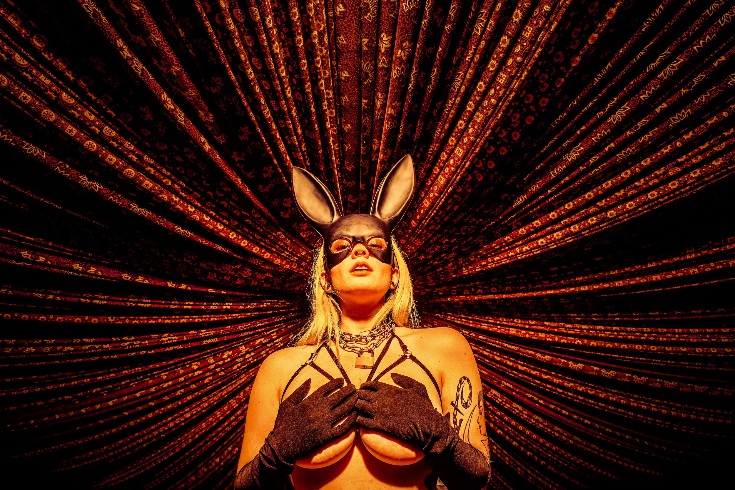 A woman with blonde hair wearing a black bunny mask and black gloves, standing in front of an ornate, red and black patterned fabric backdrop, with her hands on her chest and a chain necklace around her neck.