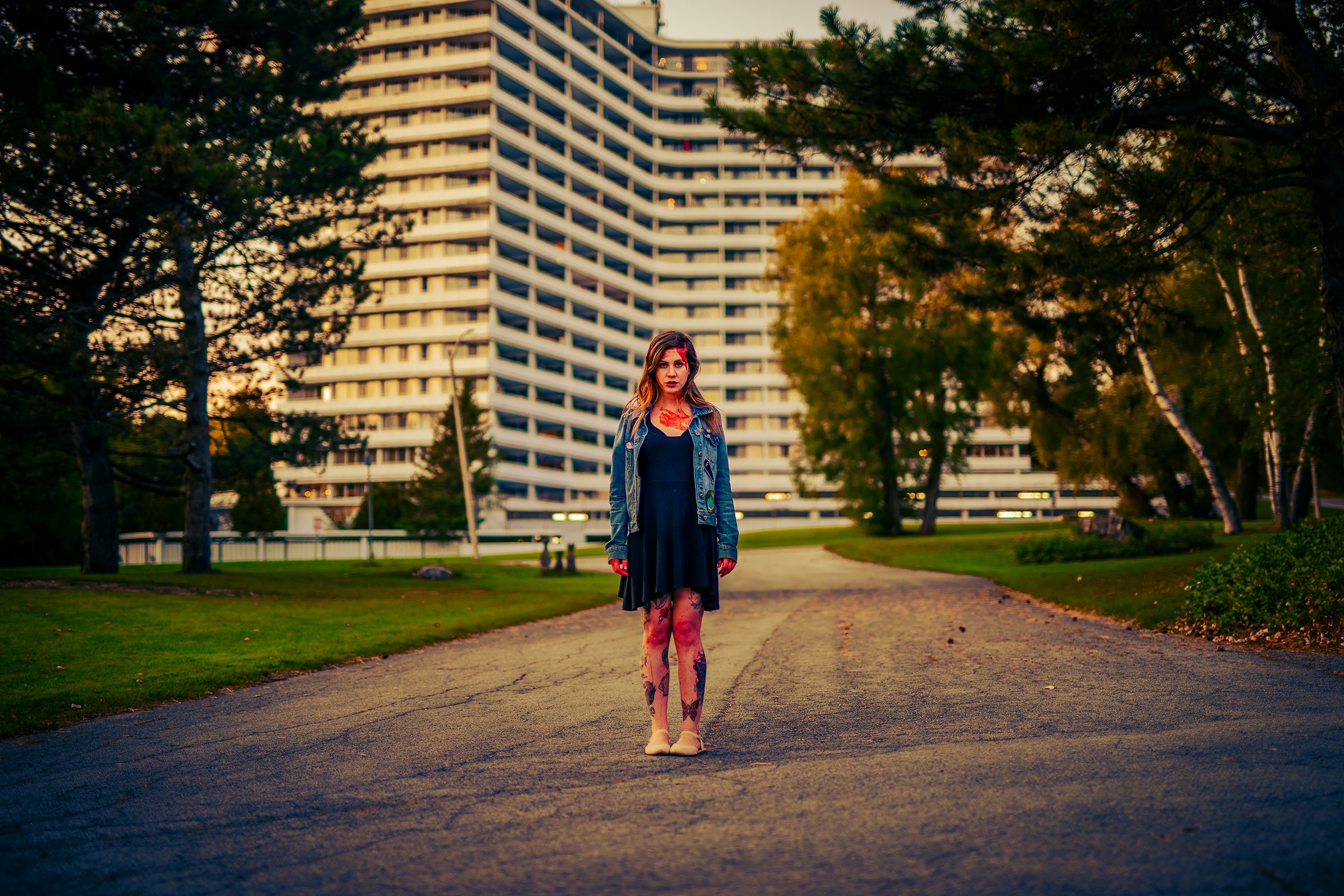 A young woman dressed in a black dress, denim jacket, and patterned tights standing on a park path with trees and a tall modern building in the background during sunset.