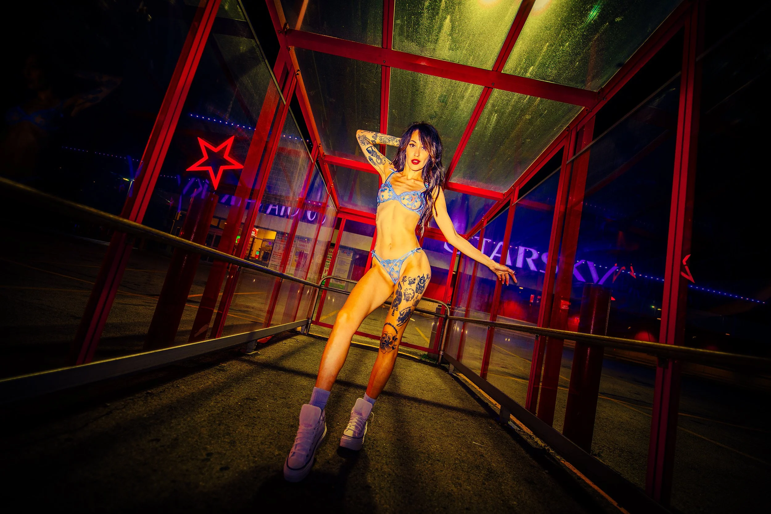 A woman with tattoos wearing a blue lace lingerie set and purple boots, standing inside a red glass structure at night with neon signs in the background.