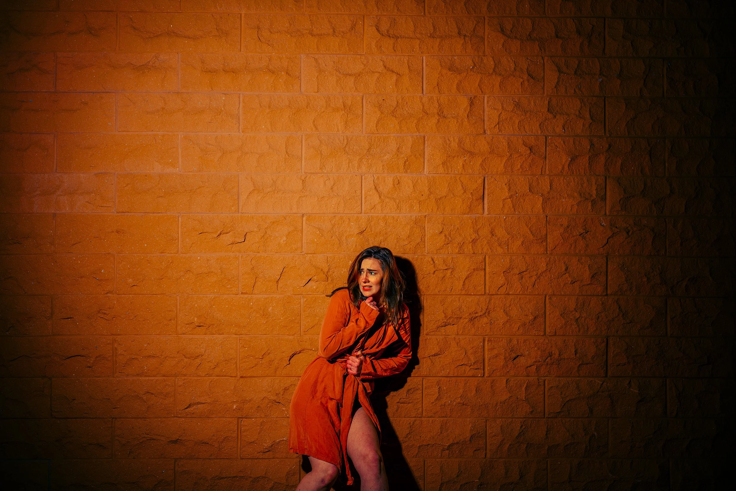 A woman in an orange dress and coat stands against a large orange brick wall, holding her coat, with a worried or distressed expression on her face.