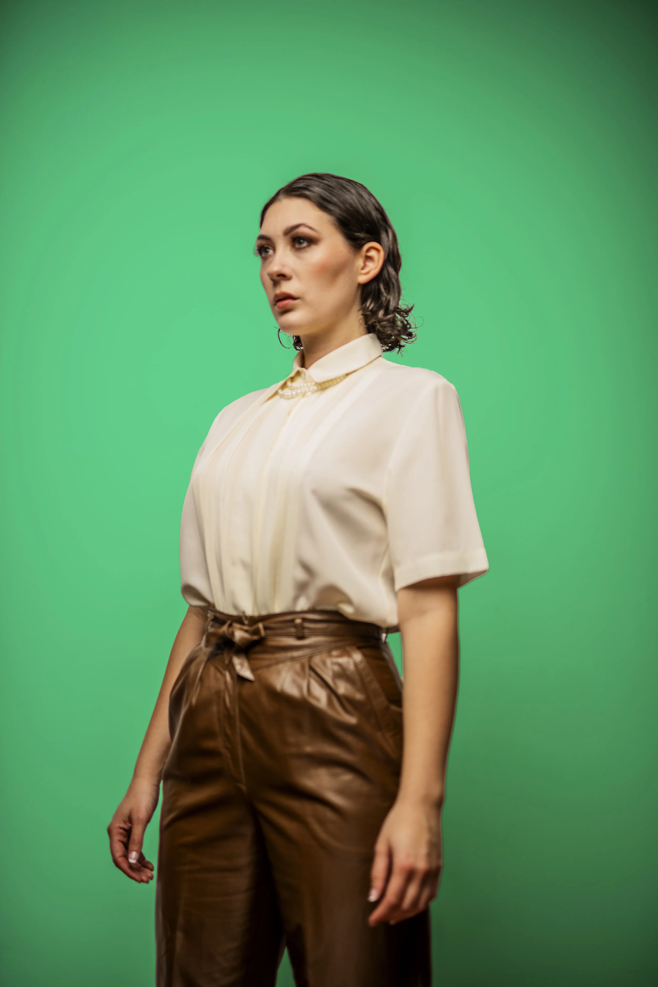 A woman with dark hair wearing a beige short-sleeve blouse and dark brown leather pants stands against a green background.