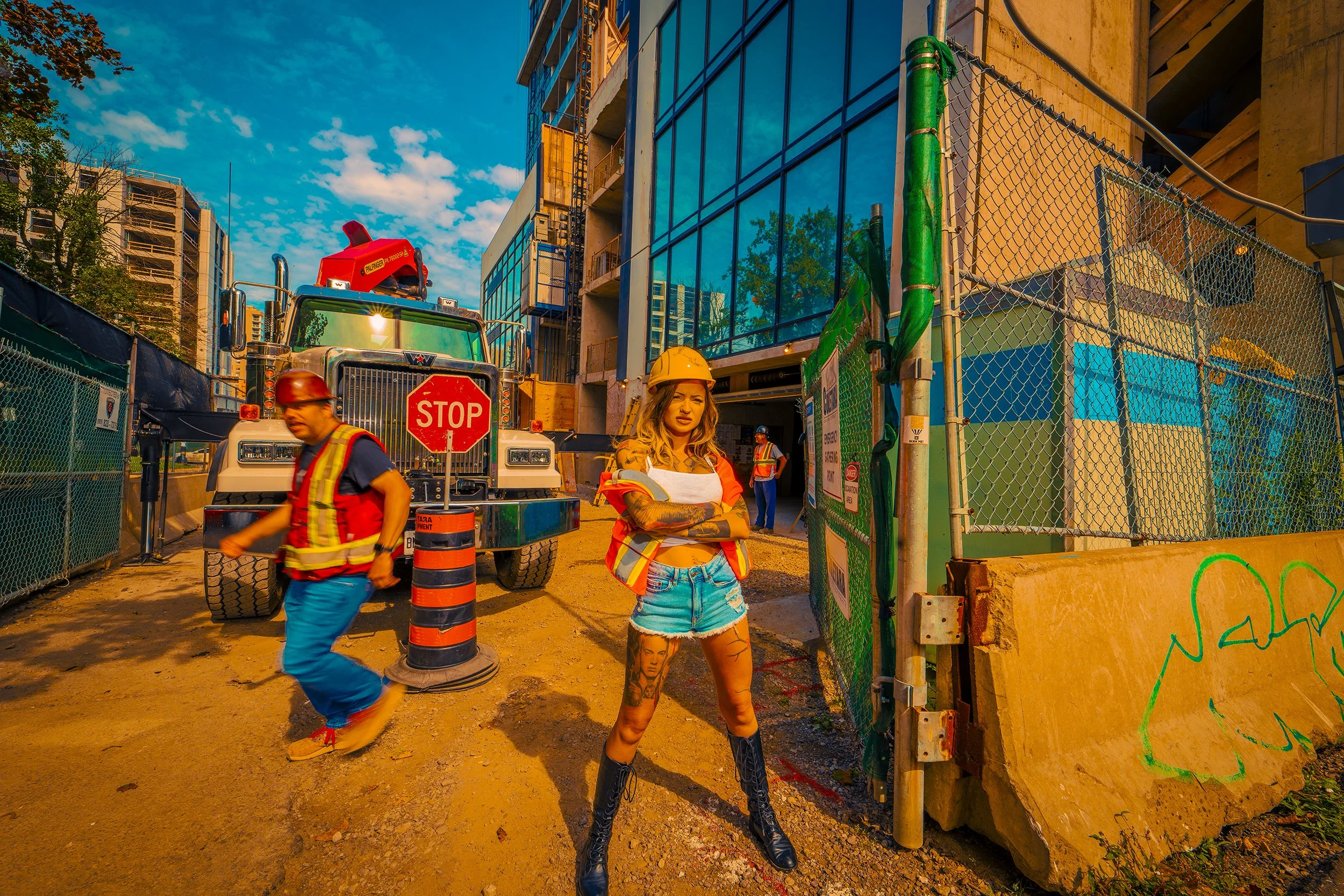 A woman with tattoos wearing a construction helmet and work vest, standing with arms crossed at a construction site, with construction workers, a stop sign, a large truck, and building structures around.