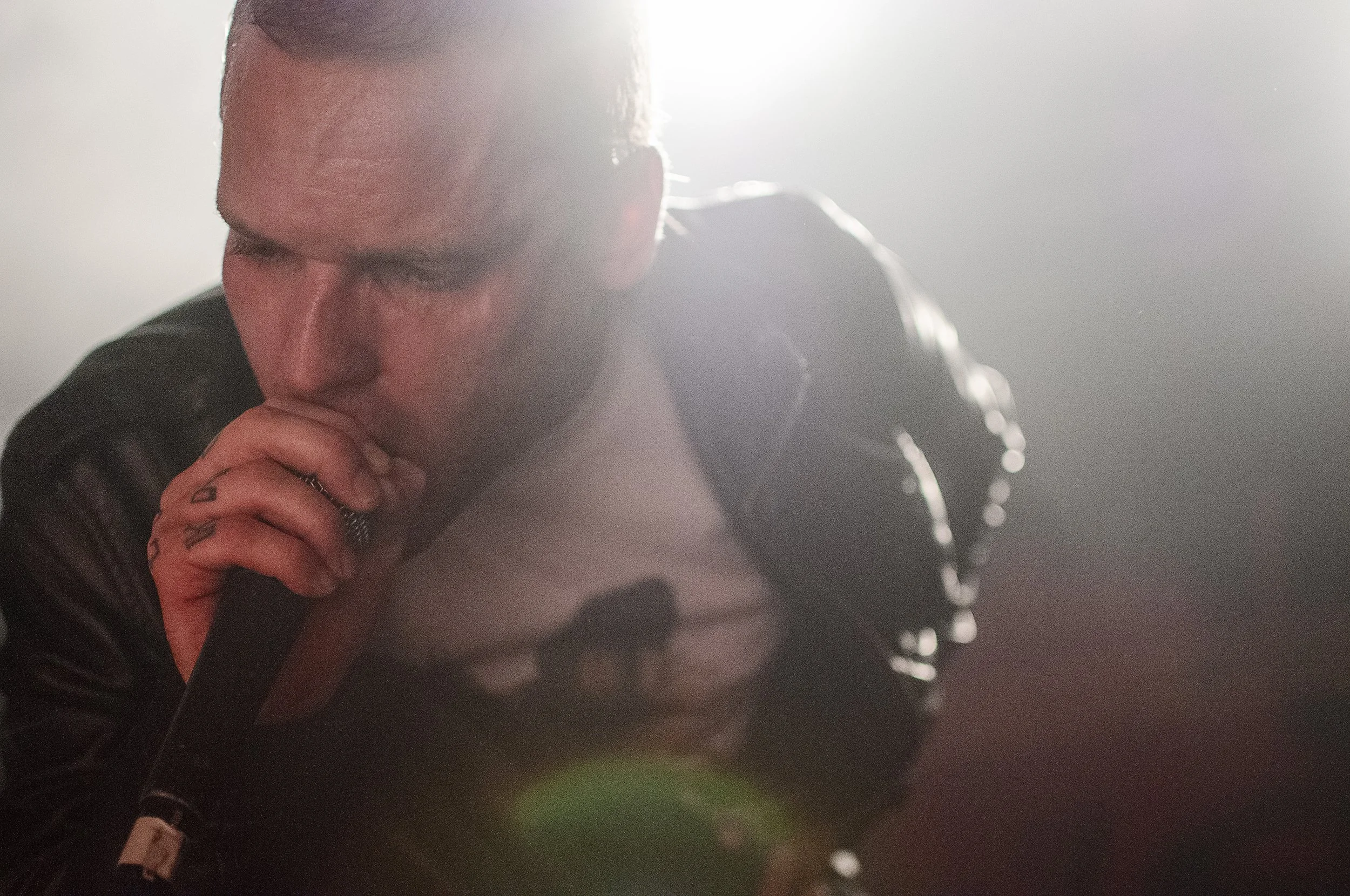 A man with closed eyes, resting his chin on his hands, wearing a black leather jacket. Bright light creates a halo effect around his head, with some water or sweat on his face, and tattoos visible on his fingers.