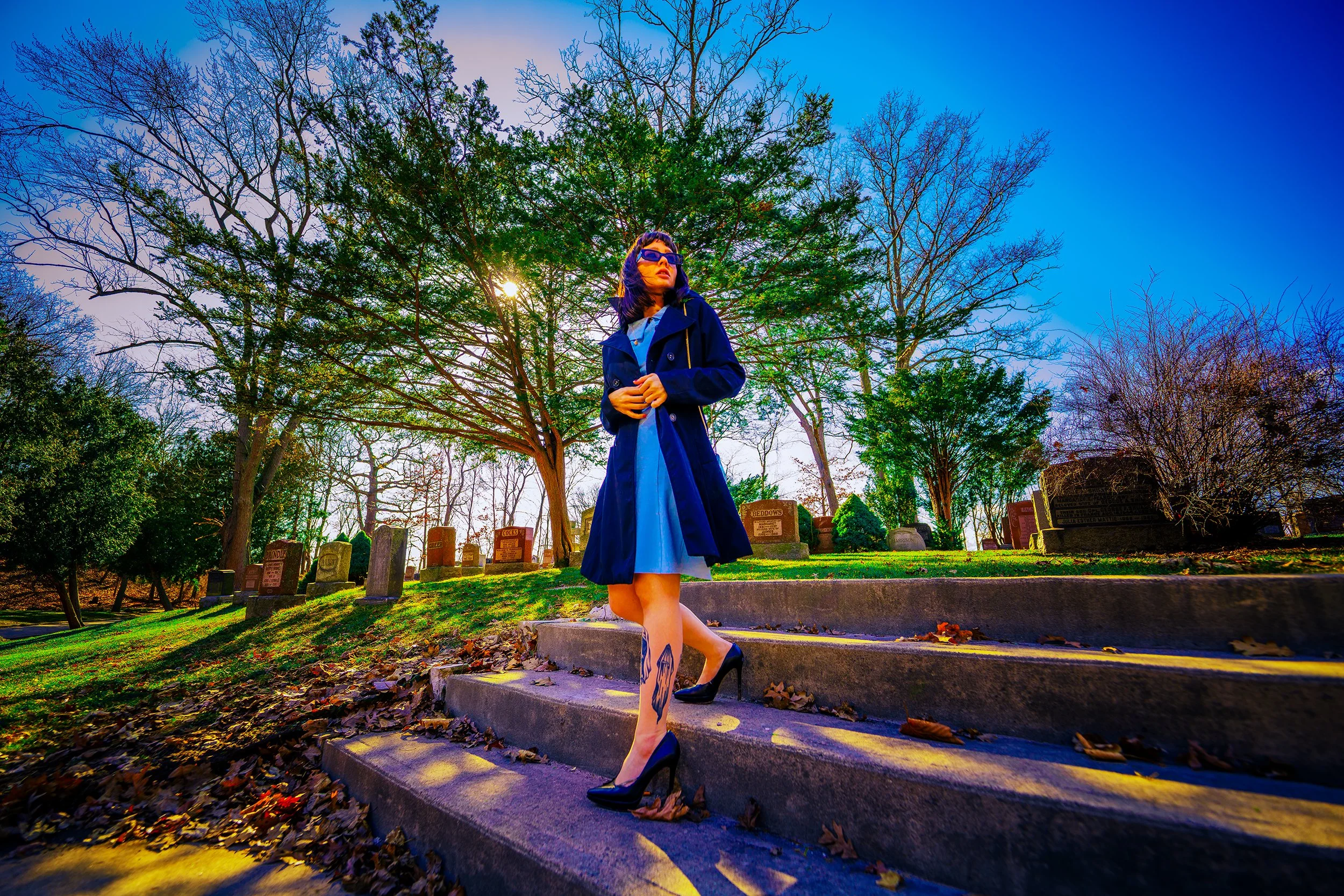 A woman wearing a navy coat, light blue dress, sunglasses, and high heels standing on steps in a cemetery with trees and headstones around, during a bright, clear day.