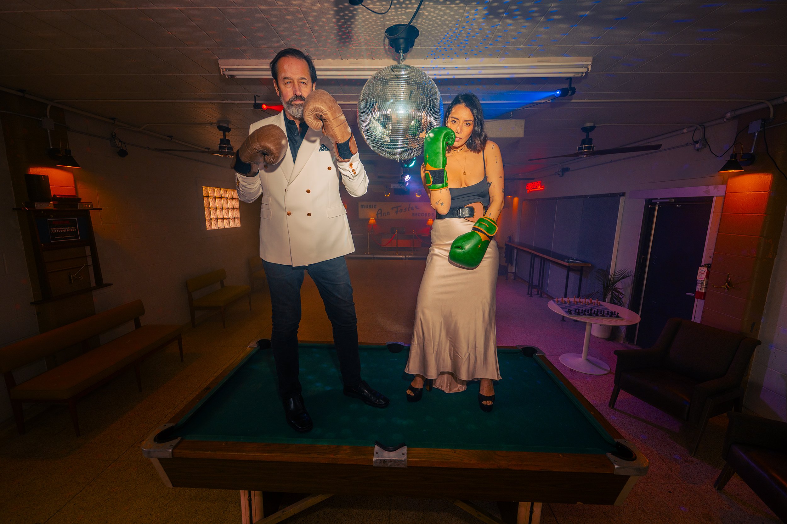 Two people standing on a pool table in a dimly lit room with disco ball, illuminated dance floor, and vintage furniture. They wear boxing gloves; the man is dressed in a white blazer and black pants, and the woman in a satin skirt and black top.