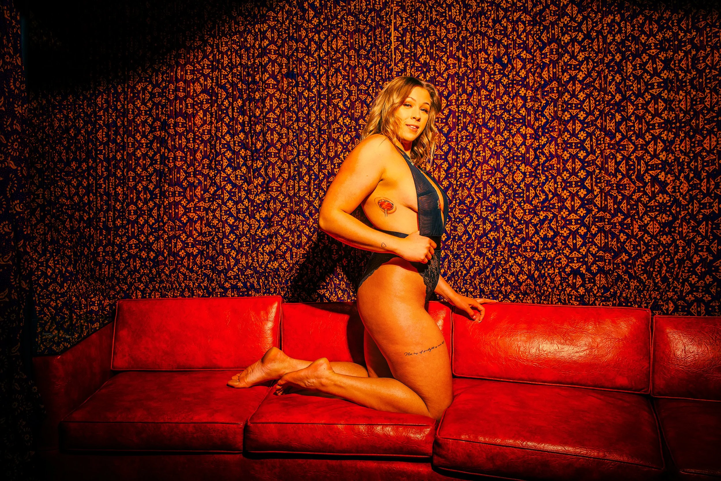 Woman with blonde hair in lingerie kneeling on a red sofa in front of a patterned orange and blue wallpaper.