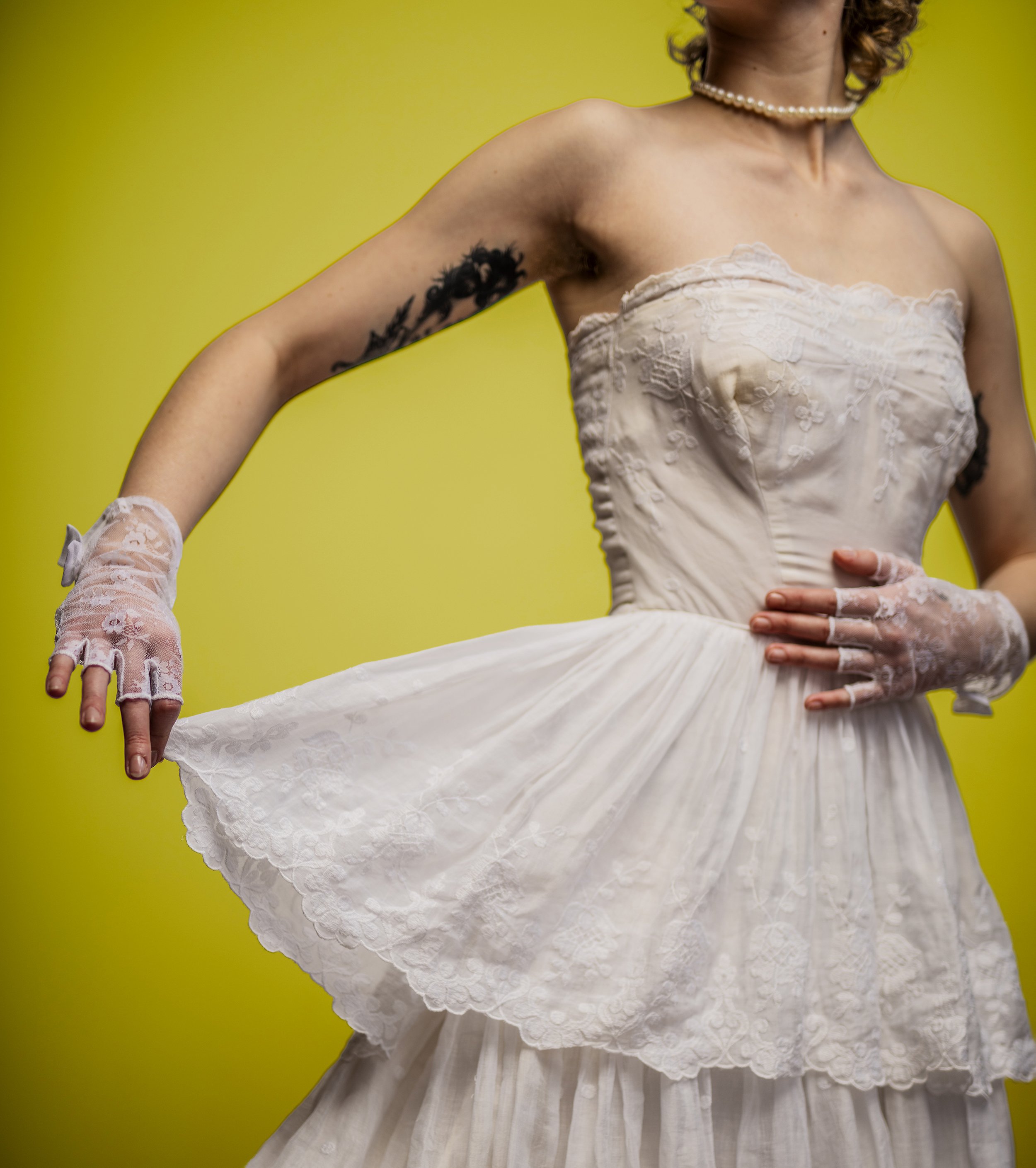 Close-up of a woman wearing a vintage white dress with lace details, a pearl necklace, lace gloves, and a black tattoo on her upper arm against a yellow background.