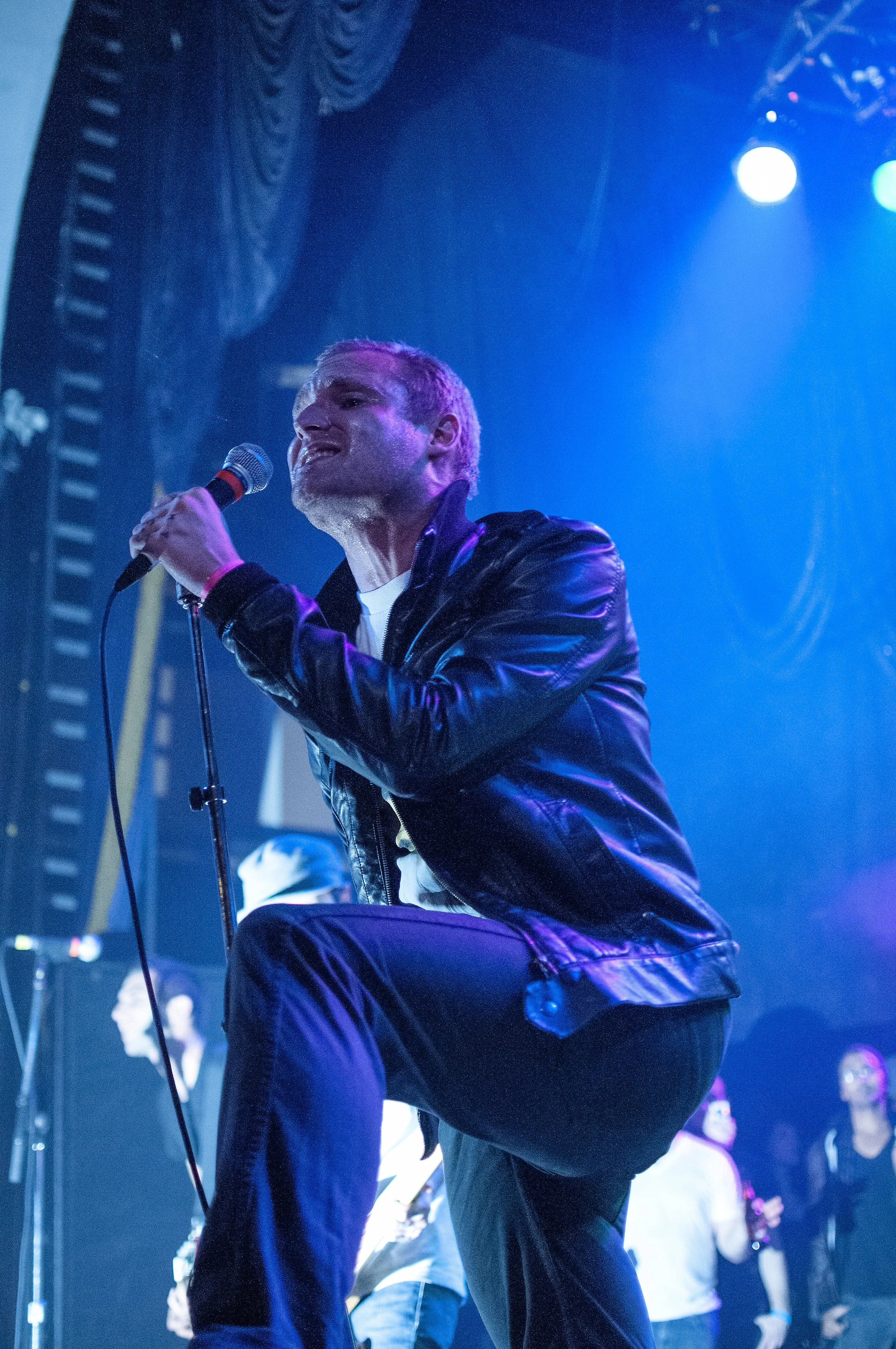 A male singer performing on stage, holding a microphone, wearing a black leather jacket and jeans, with stage lights and other people in the background.