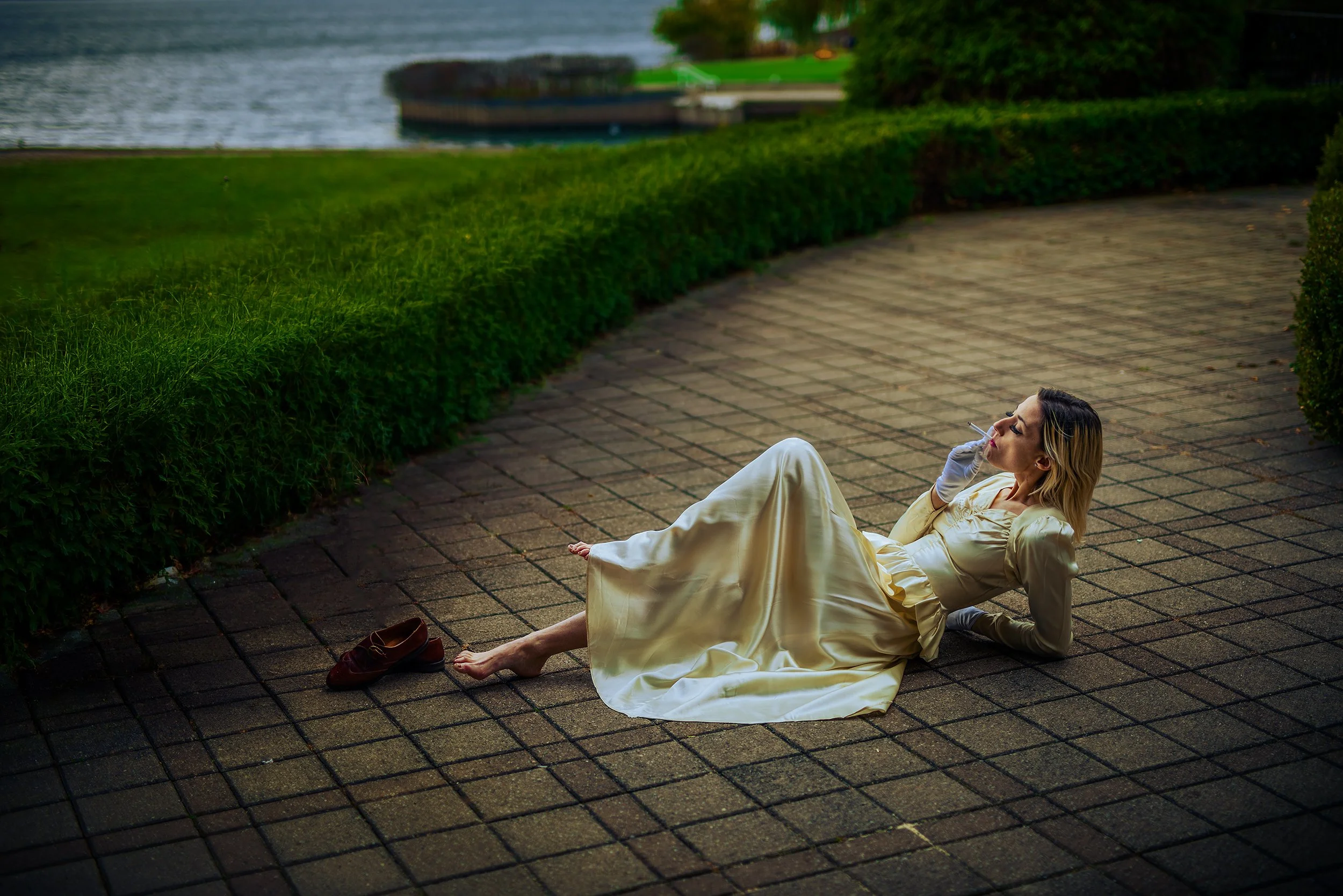 Woman in a white satin dress and gloves sitting on a brick pathway by a hedge, smoking a cigarette near a river, with a cityscape in the background.