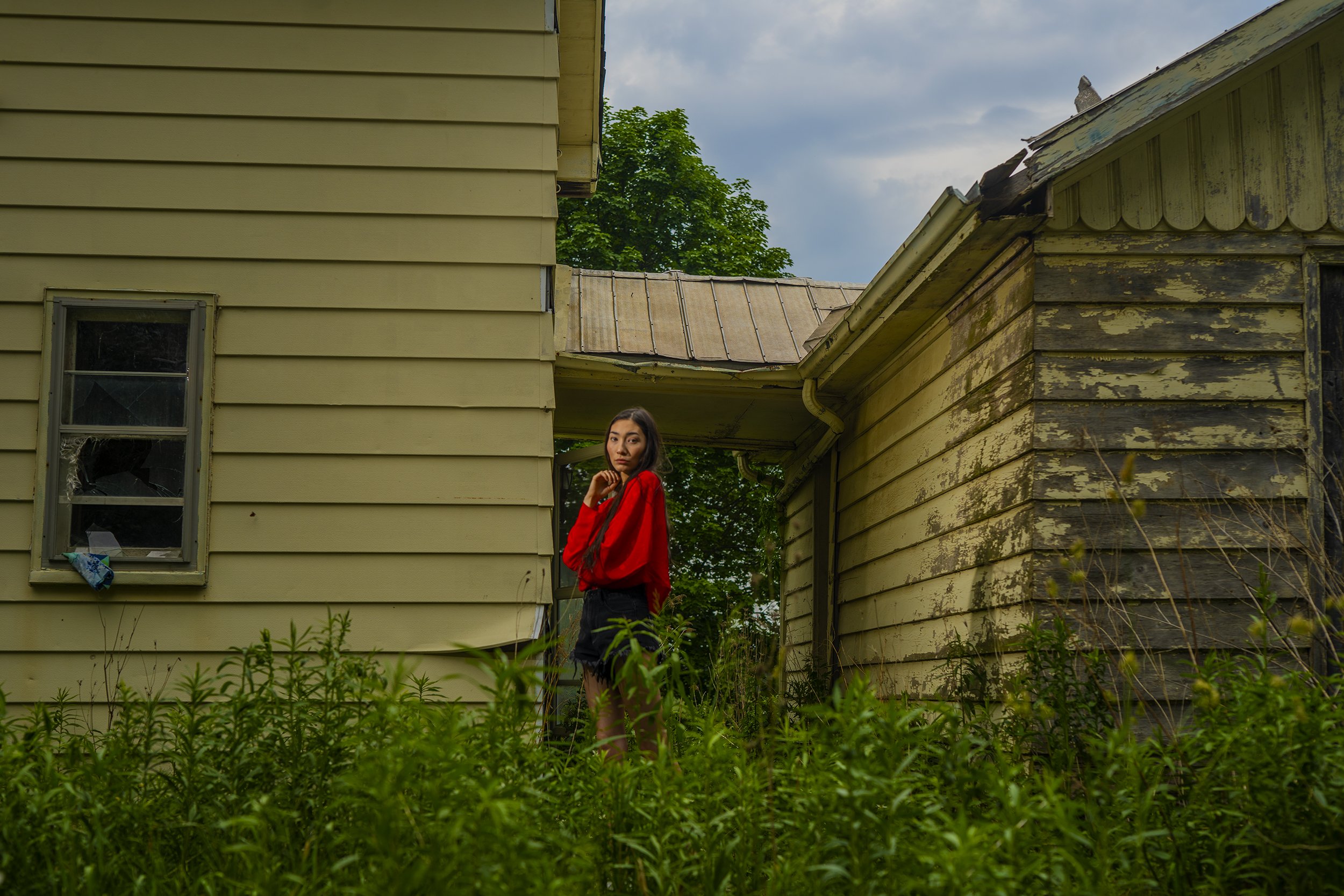 A young woman with long dark hair wearing a red jacket and black shorts stands outside between two weathered, yellow-painted houses with peeling paint and damaged roofs. She is looking at the camera with a serious expression, and there are green plan
