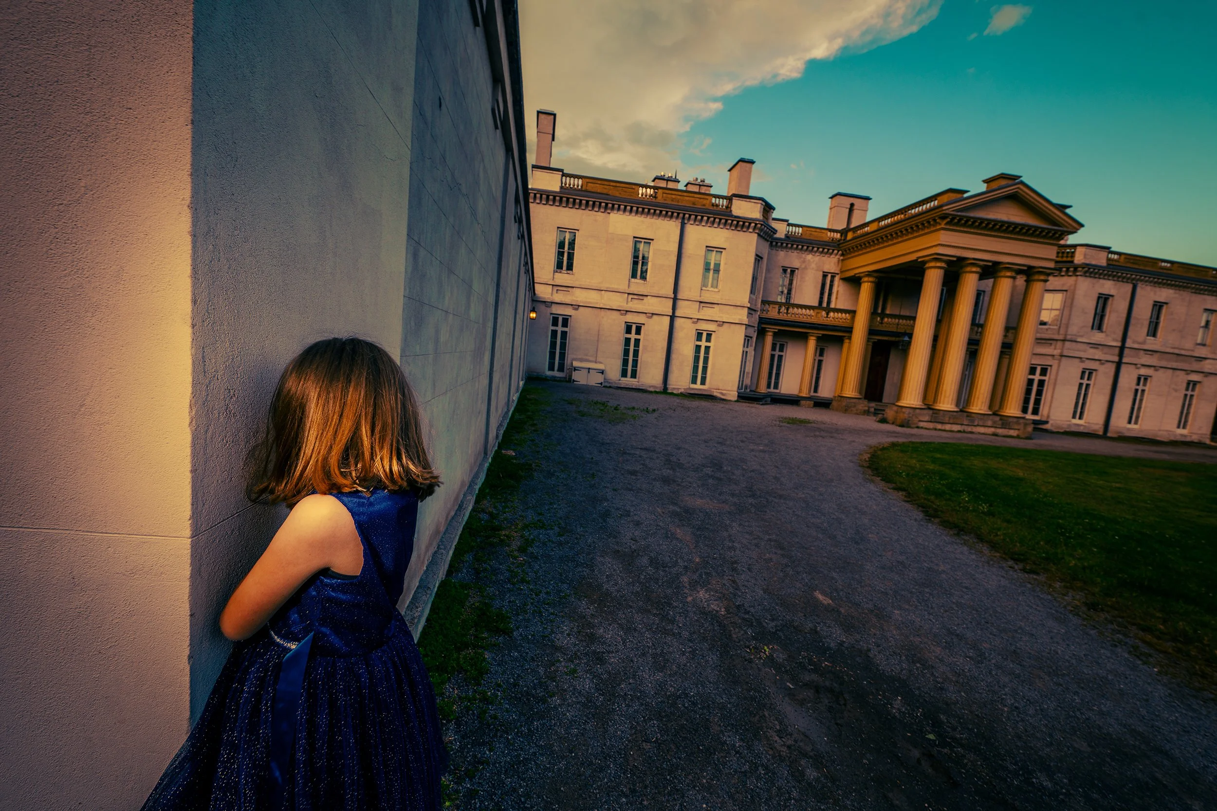 A young girl with brown hair, wearing a blue dress, stands next to a wall outside a large historic building with columns and a portico, during sunset.