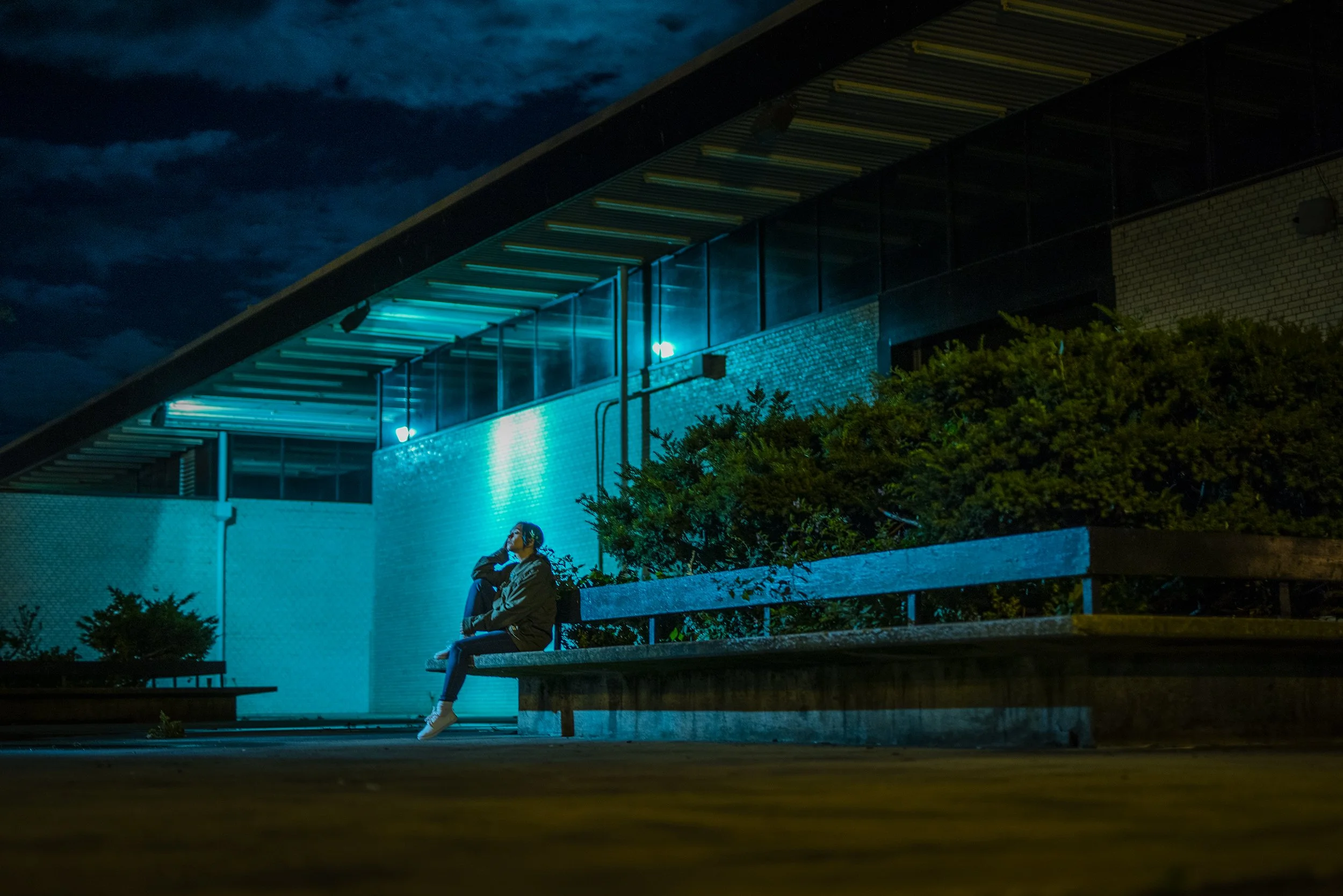 A person sitting alone on a park bench at night, wearing headphones, in front of a modern building with blue lighting and a cloudy sky.