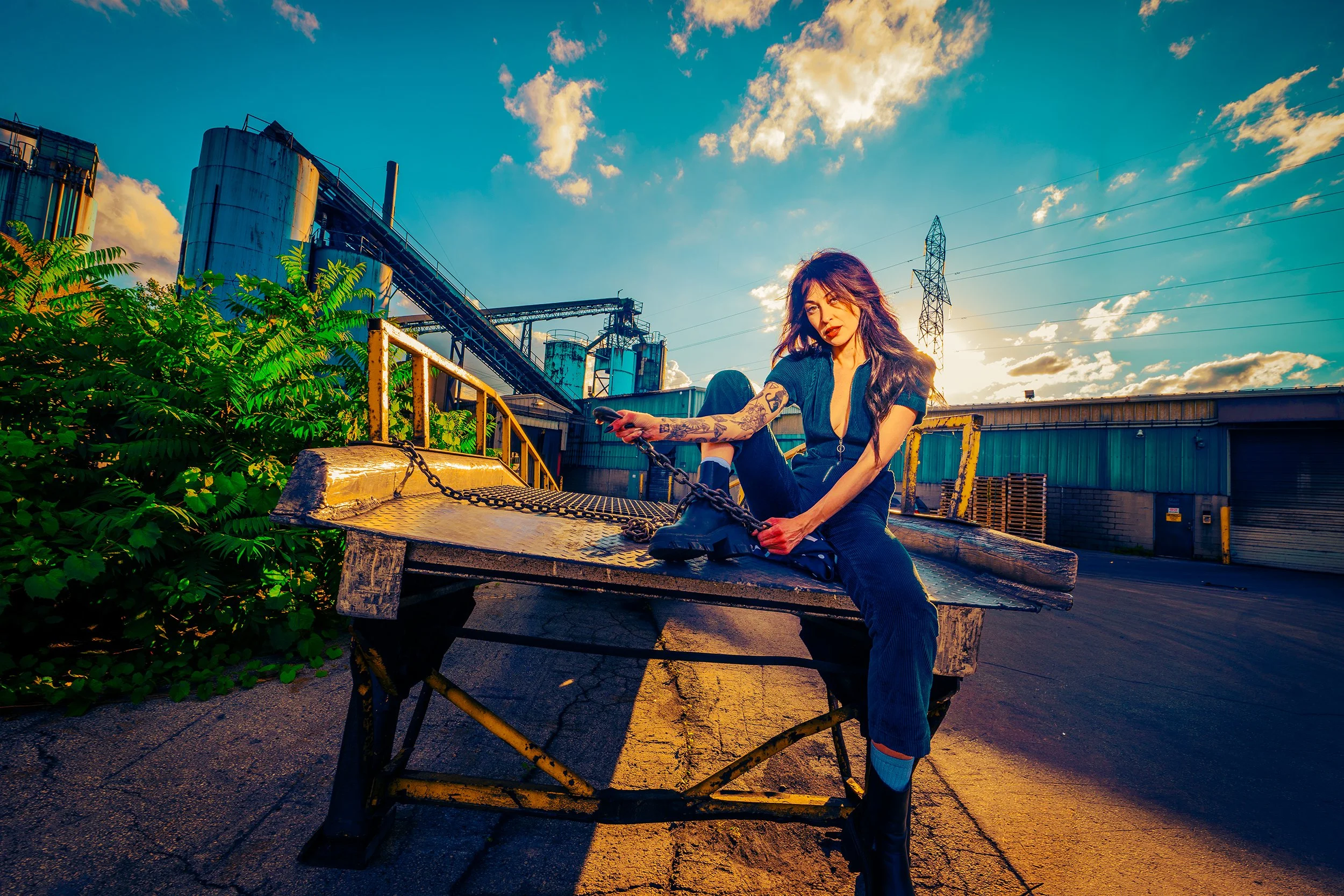 A woman with tattoos on her arms is sitting on an old metal coal cart in an industrial outdoor setting during sunset, holding a shoe and looking at the camera. There are industrial silos, a conveyor belt, power lines, and a building in the background