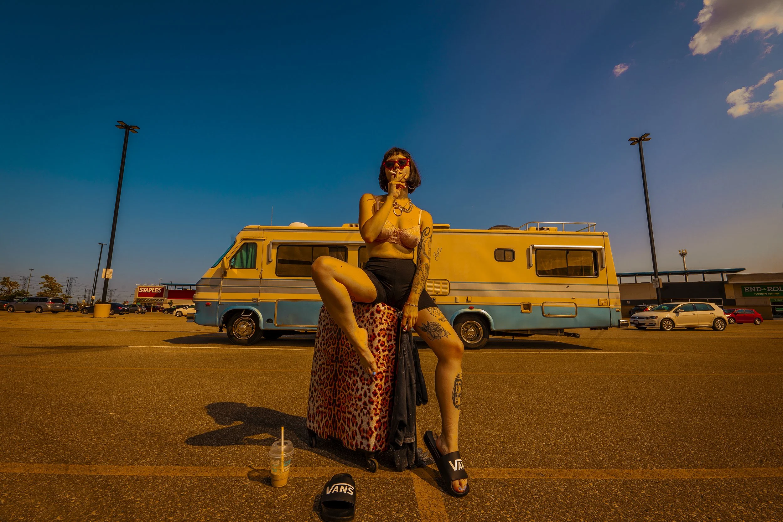 A woman wearing sunglasses and a bra top, with tattoos, sitting on a leopard print ottoman in a parking lot with a yellow and blue RV in the background, and a Starbucks drink and black Vans slides on the ground.