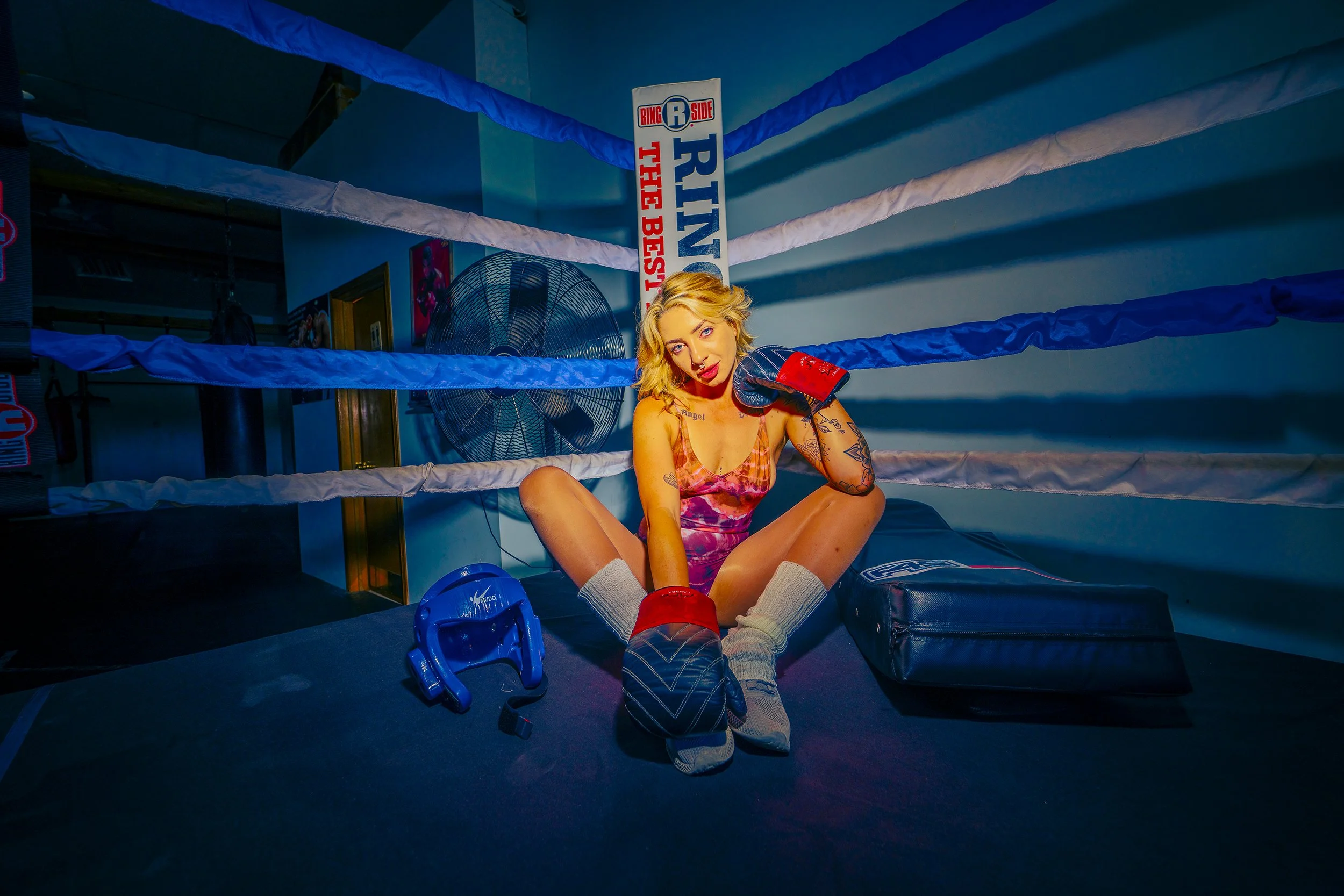 A woman sits inside a boxing ring, wearing boxing gloves and a colorful tank top, with boxing shoes and gray socks on. She has blonde hair and tattoos on her arms. A large fan is visible in the background, along with a sign that reads "Ring" and "The