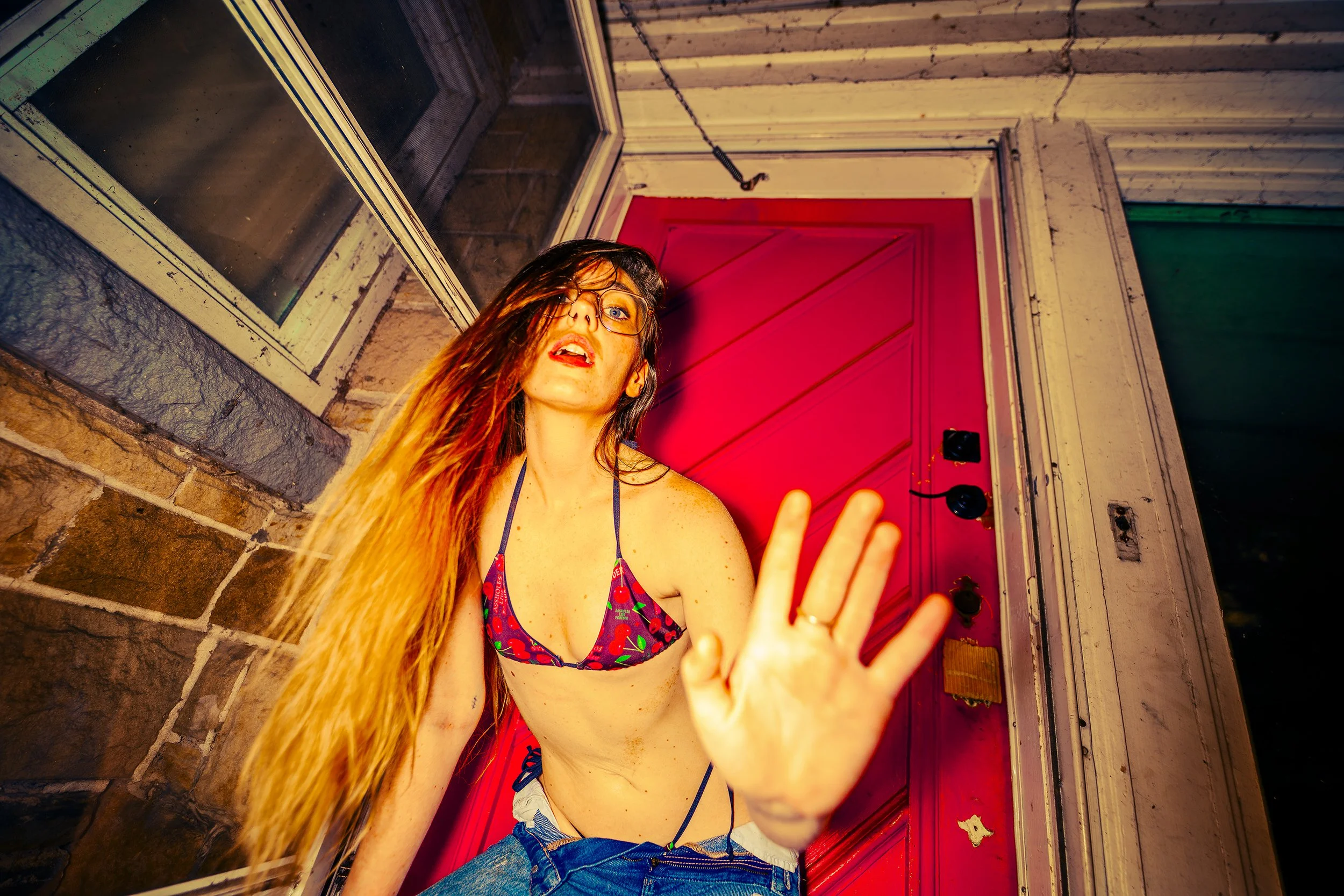 A woman with long hair, glasses, and a colorful patterned bikini top is reaching towards the camera with her hand, blocking part of her face. She is standing outside near a brick wall and a red door, with a window and a speaker nearby.
