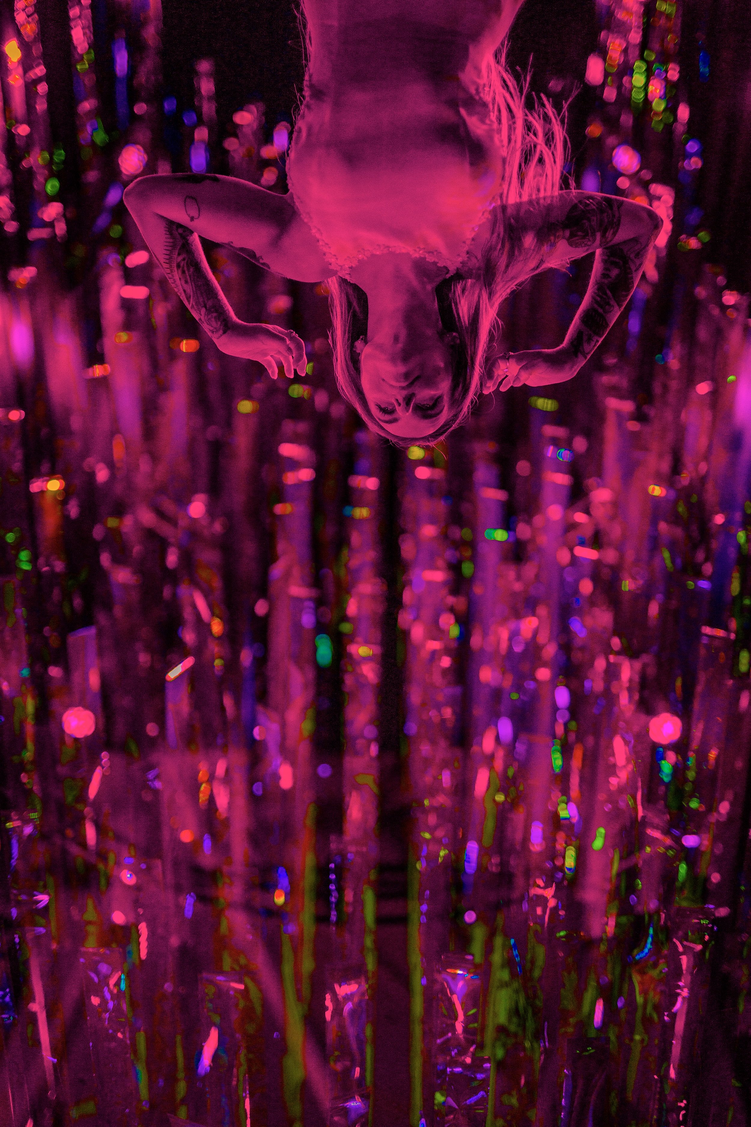 A woman with tattoos on her arms and long hair is posing with her hands behind her head in front of vibrant, colorful, shimmering background lights.