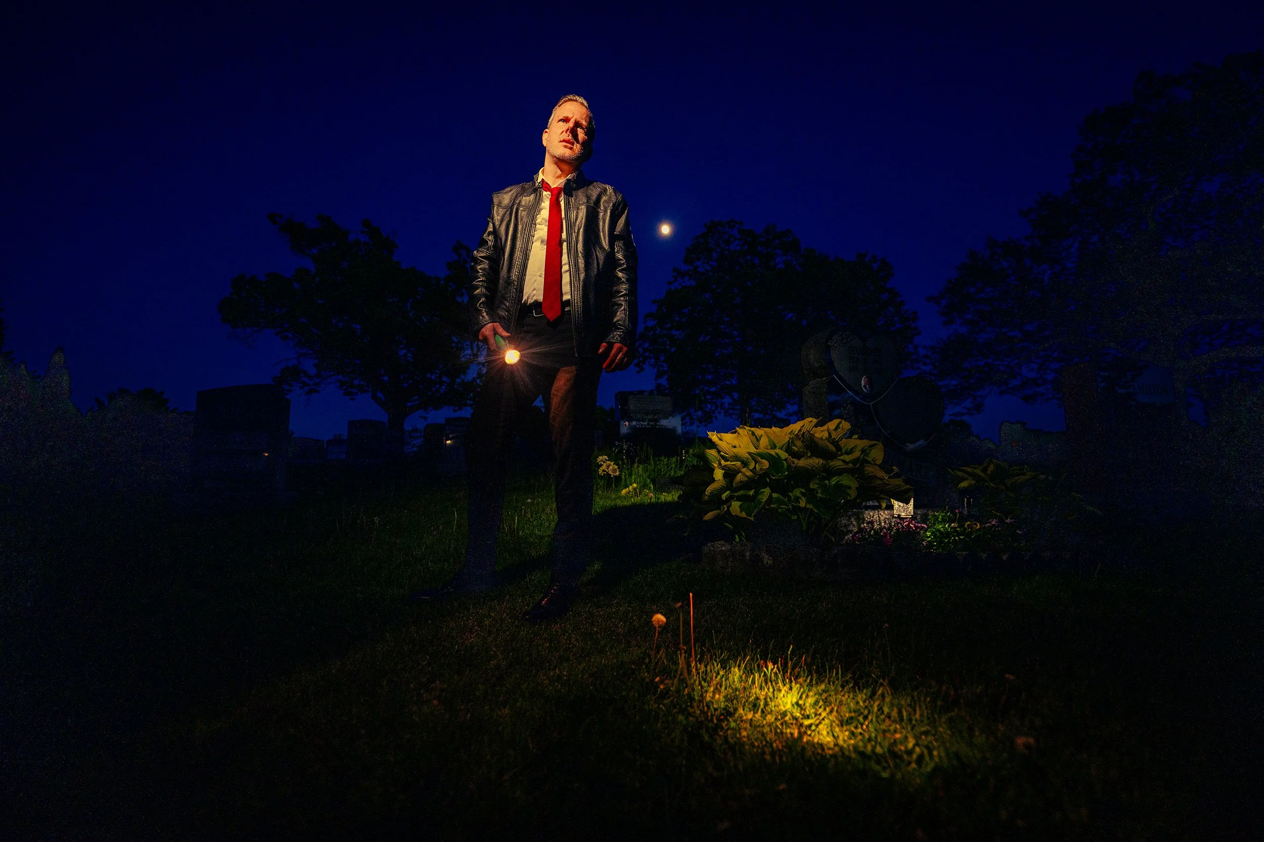 A man standing in a graveyard at night, holding a flashlight, wearing a leather jacket, white shirt, and red tie, with the moon in the dark sky behind him, surrounded by trees and tombstones.
