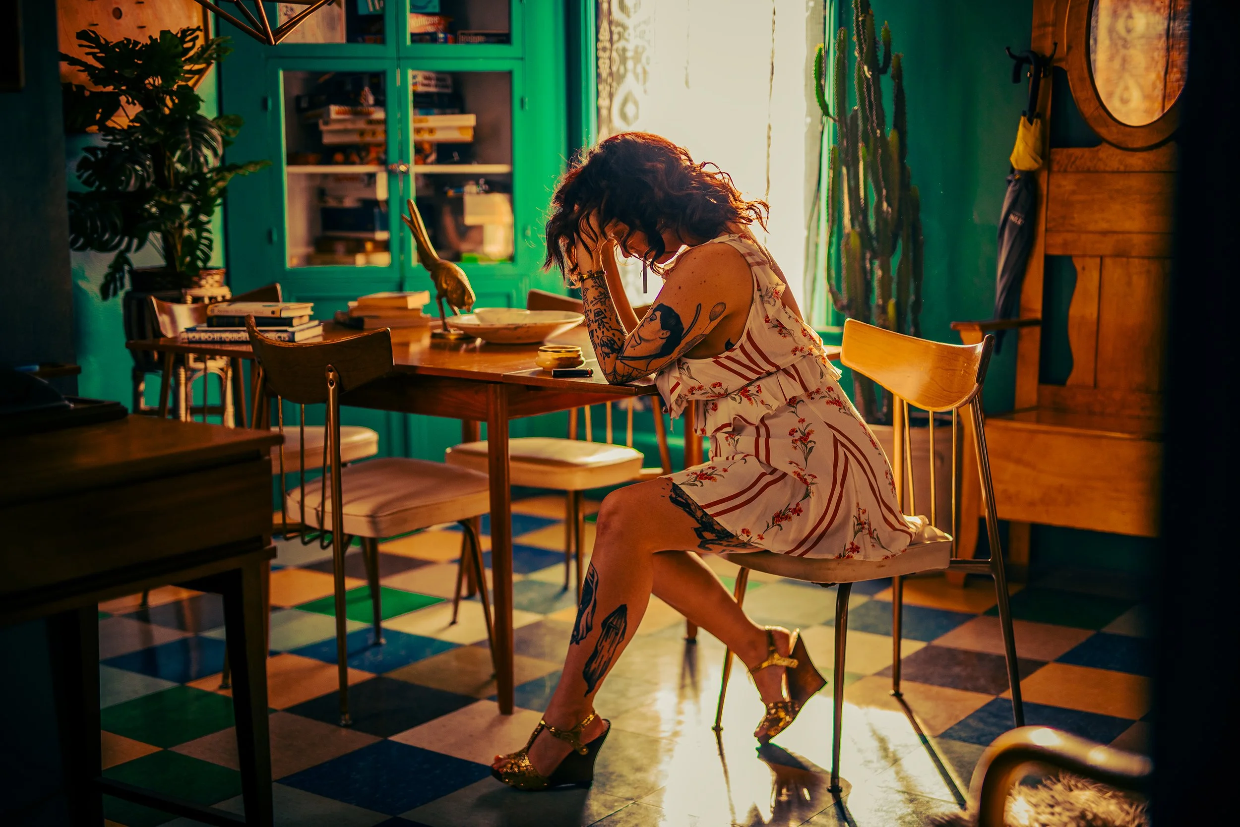 A woman with tattoos and curly hair in a floral dress sitting at a dining table, looking downward, with her hand on her forehead, in a colorful room with green cabinets, plants, books, and a checkered floor.