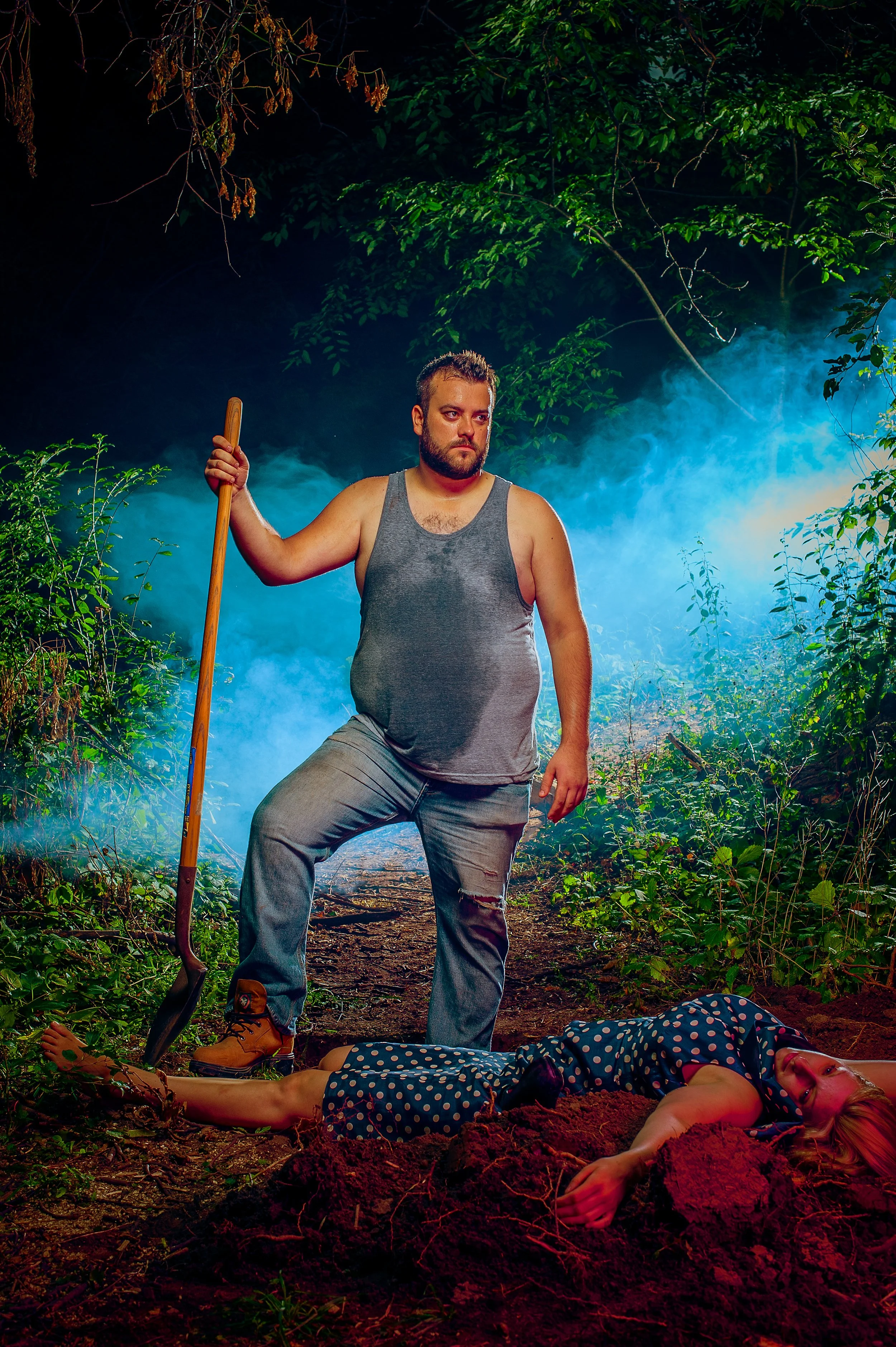 A man with a beard and a gray tank top stands in a forest at night, holding a shovel and looking down at a woman lying on the ground who appears to be dead, wearing a blue dress with white polka dots. There is smoke or fog in the background, creating