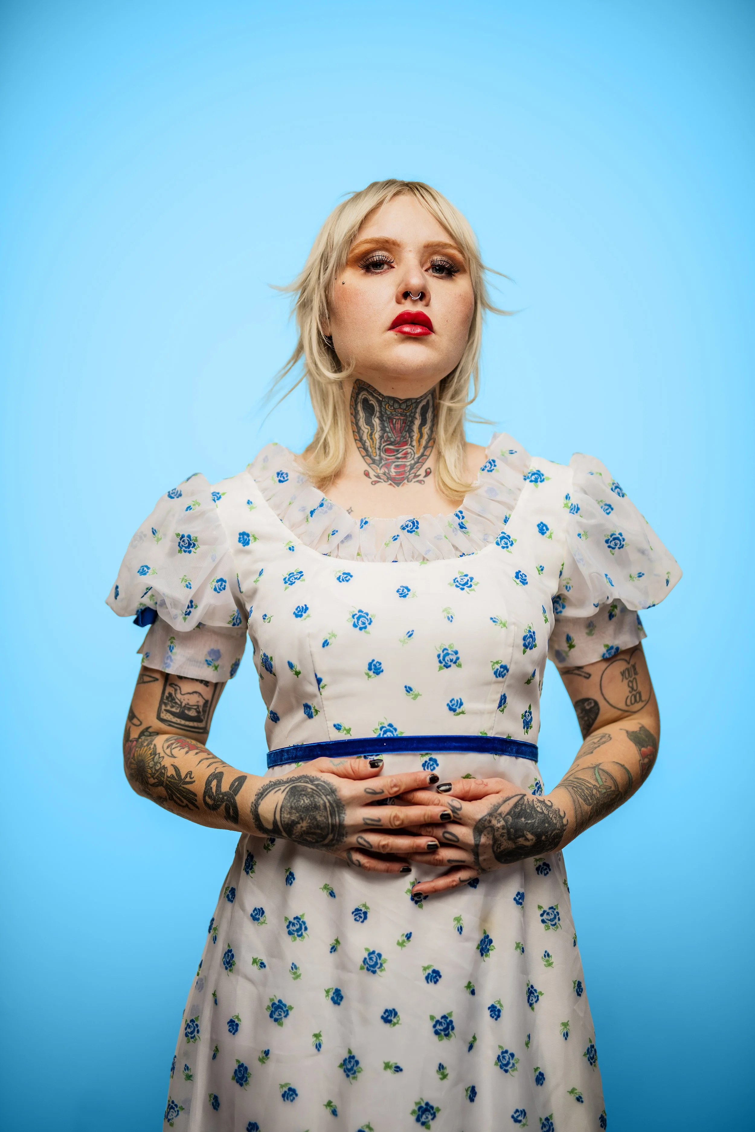 A woman with blonde hair and tattoos wearing a white dress with blue floral pattern, standing against a bright blue background, looking directly at the camera.