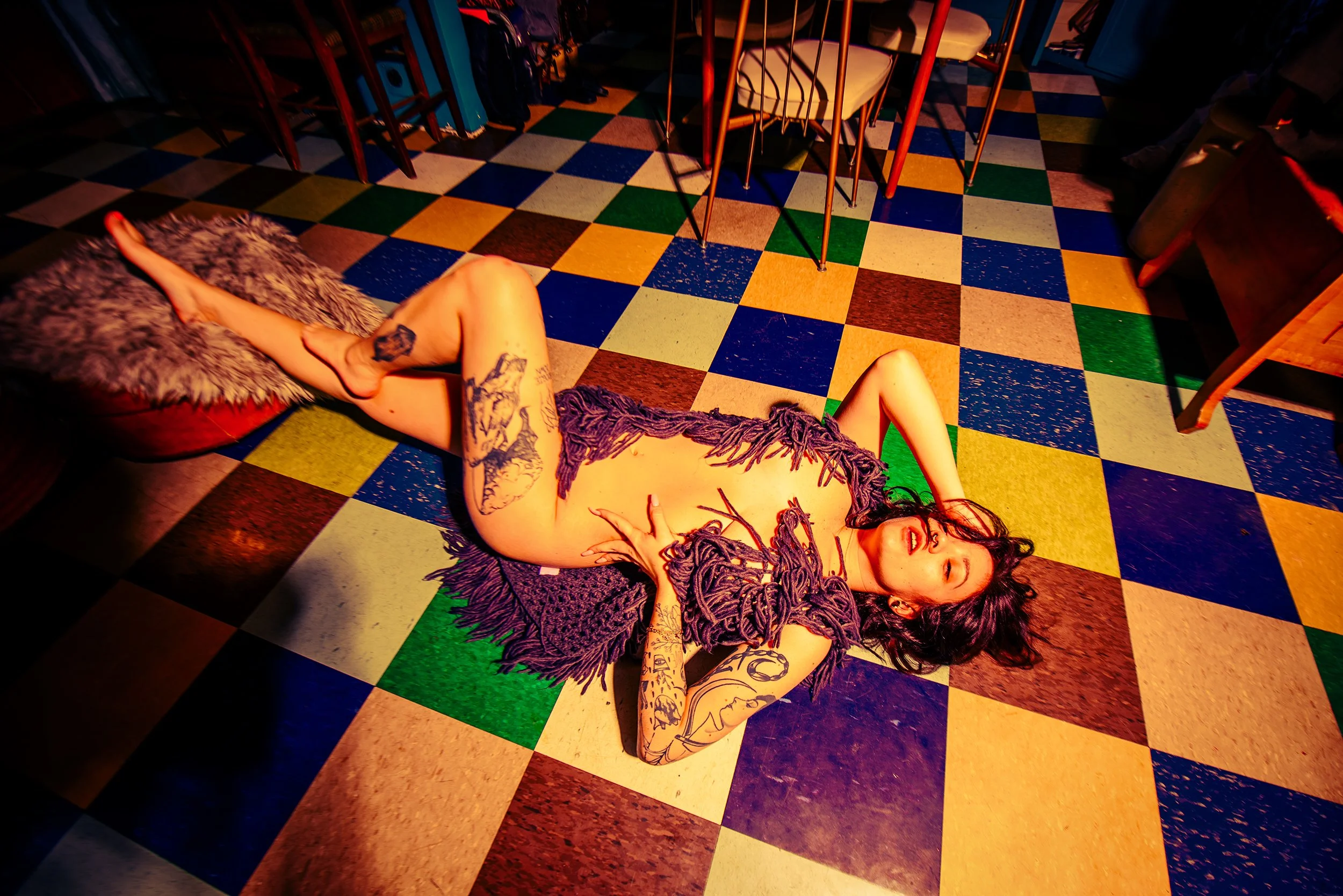 A woman with tattoos and long dreadlocks lying on a colorful checkered floor in a dimly lit room, surrounded by chairs and furniture.