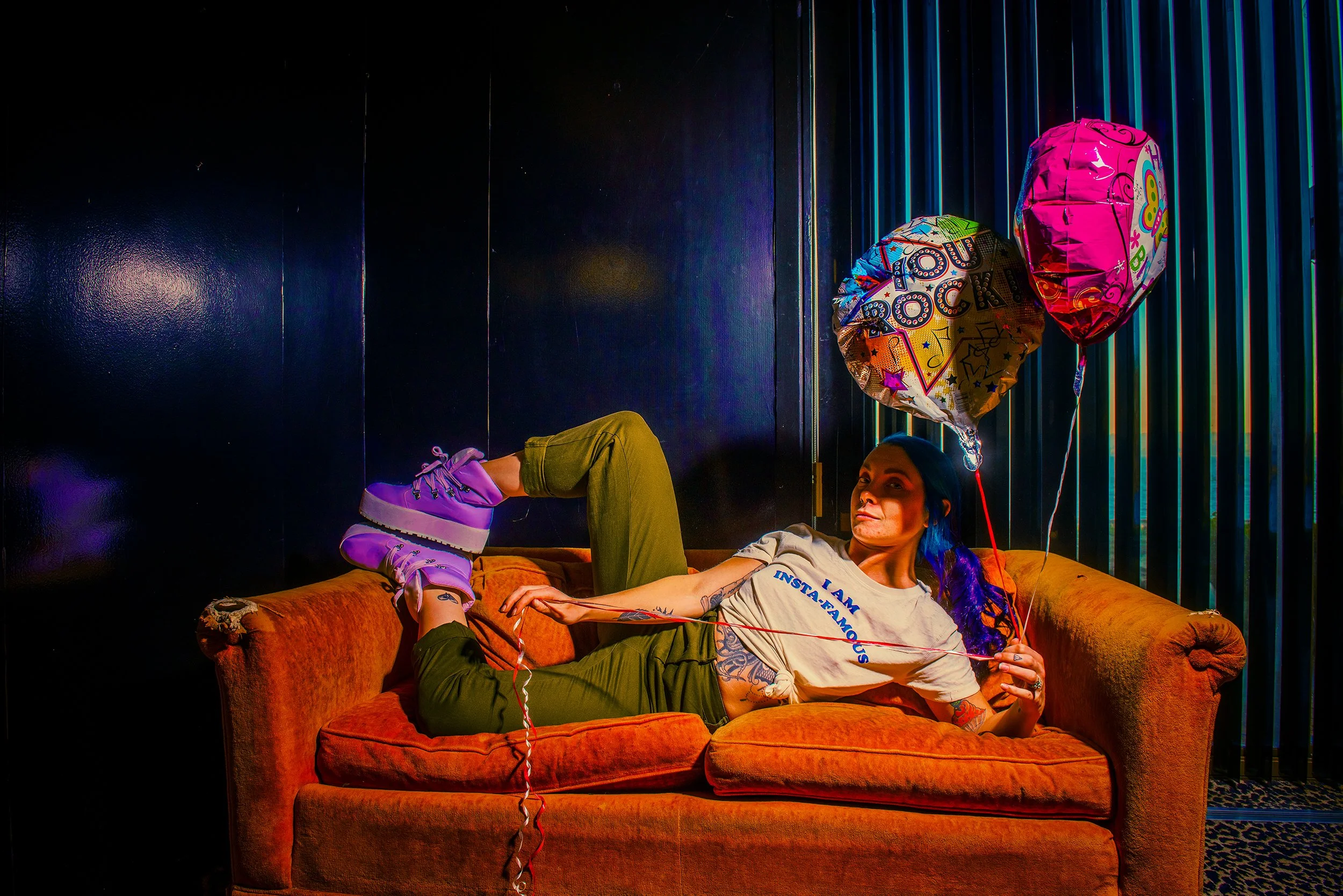A woman with tattoos wearing a headscarf and a T-shirt sitting on an orange velvet couch, holding balloons with bright colors and decorative designs, in a dark room with colorful vertical light stripes on the wall.