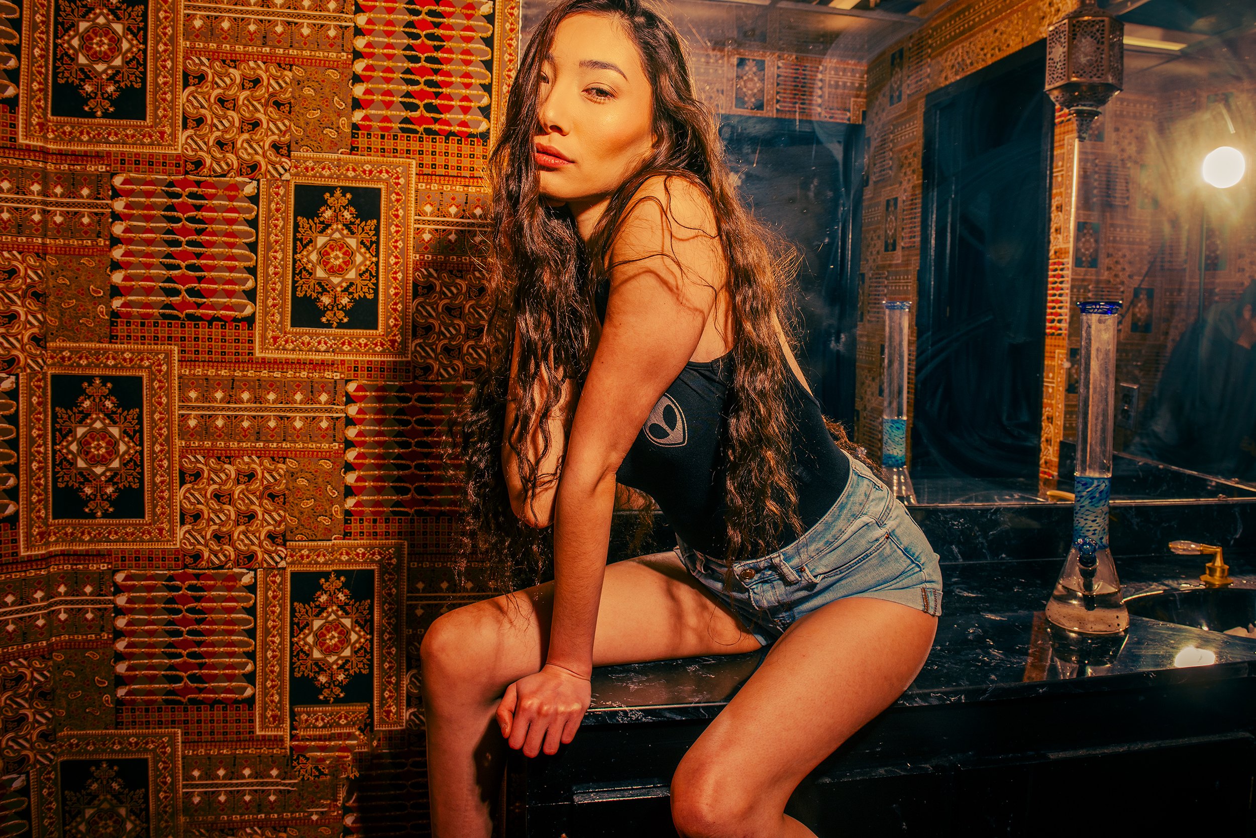 A young woman with long wavy hair in a black tank top and denim shorts is kneeling on a black marble surface, posing confidently with a colorful patterned wall in the background and warm lighting.