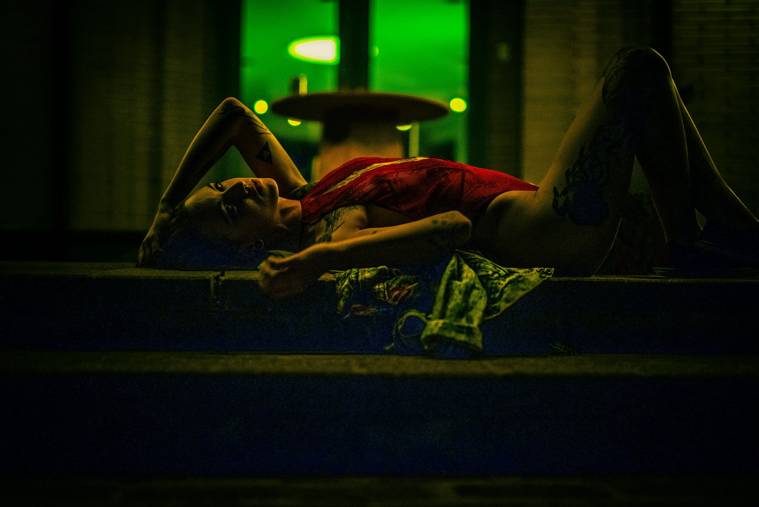 A woman with tattoos lying on her back on a dark floor, wearing a red dress, in a room with green lighting