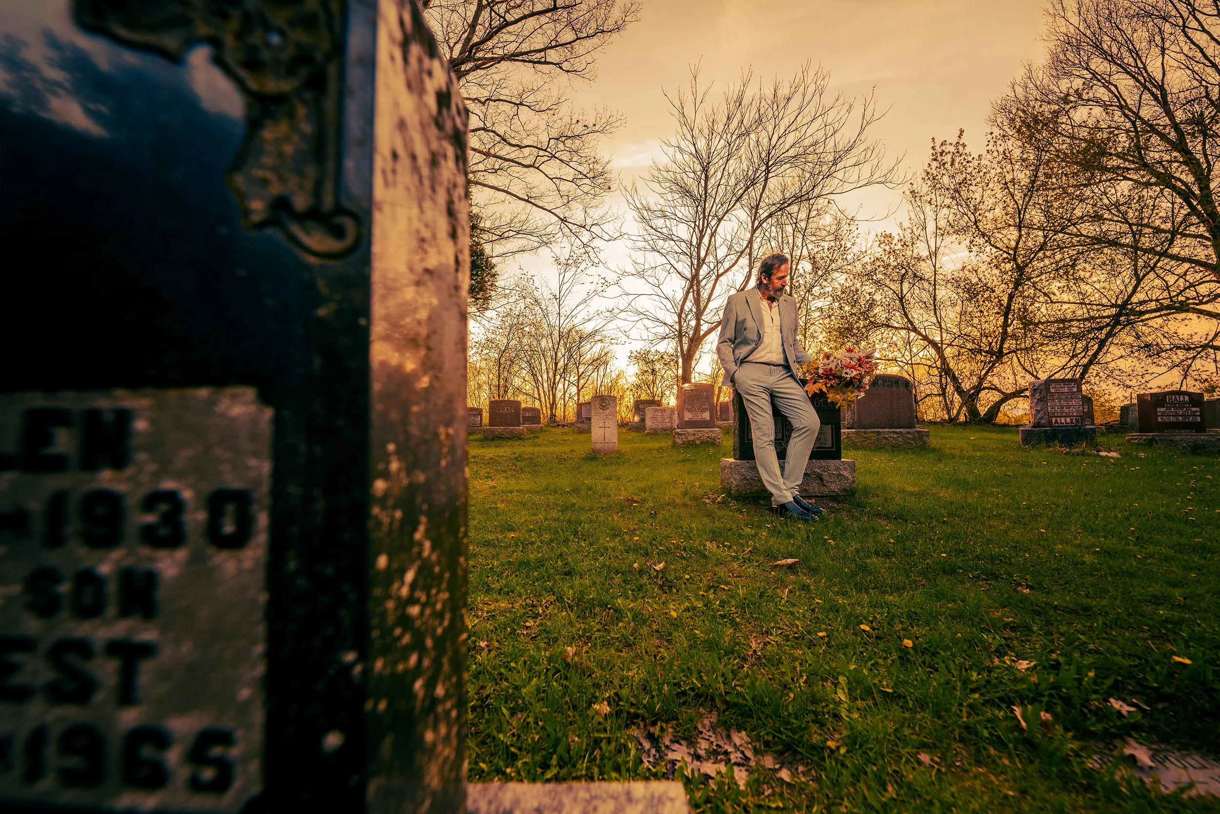 A man in a light gray suit leaning against a gravestone decorated with flowers in a cemetery during sunset, with leafless trees in the background.