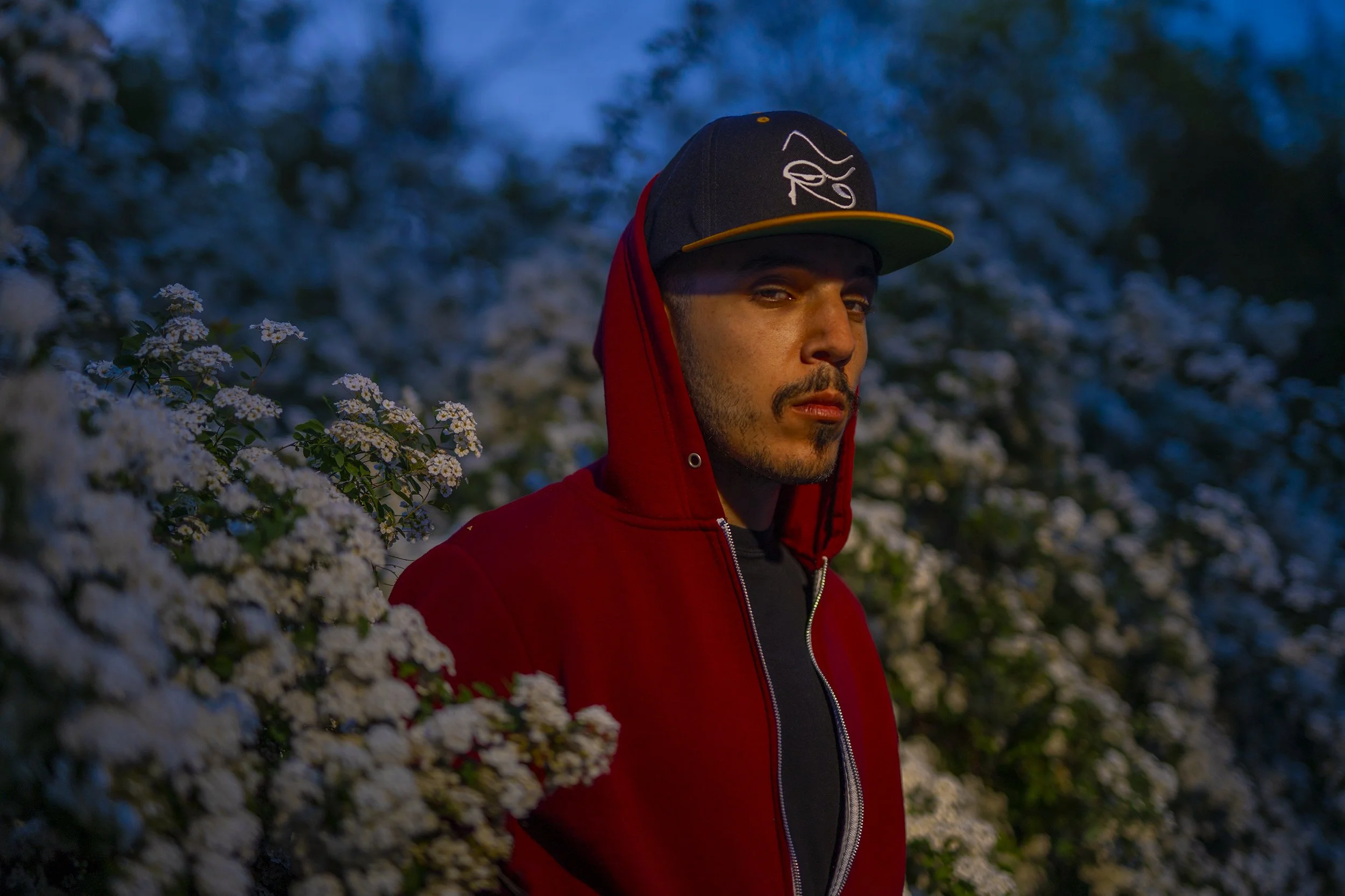 A young man with a beard wearing a black and yellow baseball cap and a red hoodie, standing outdoors near white flowering bushes during dusk or early evening.