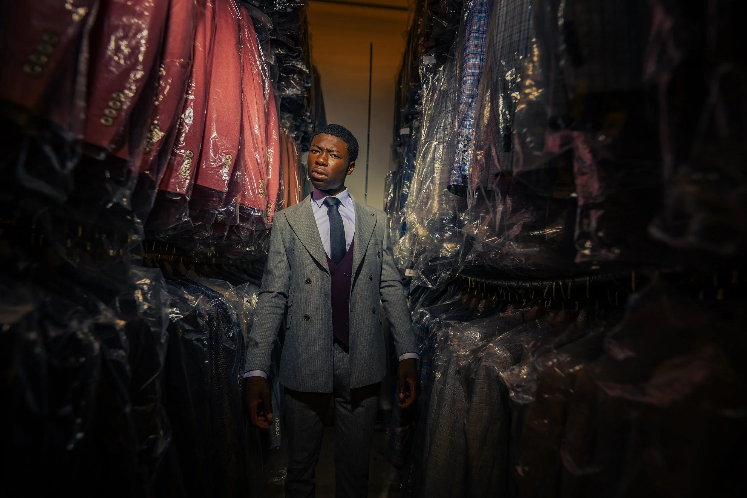 A young man in a gray suit with a purple vest and tie looking confused in a closet filled with hanging suits and jackets covered in plastic bags.