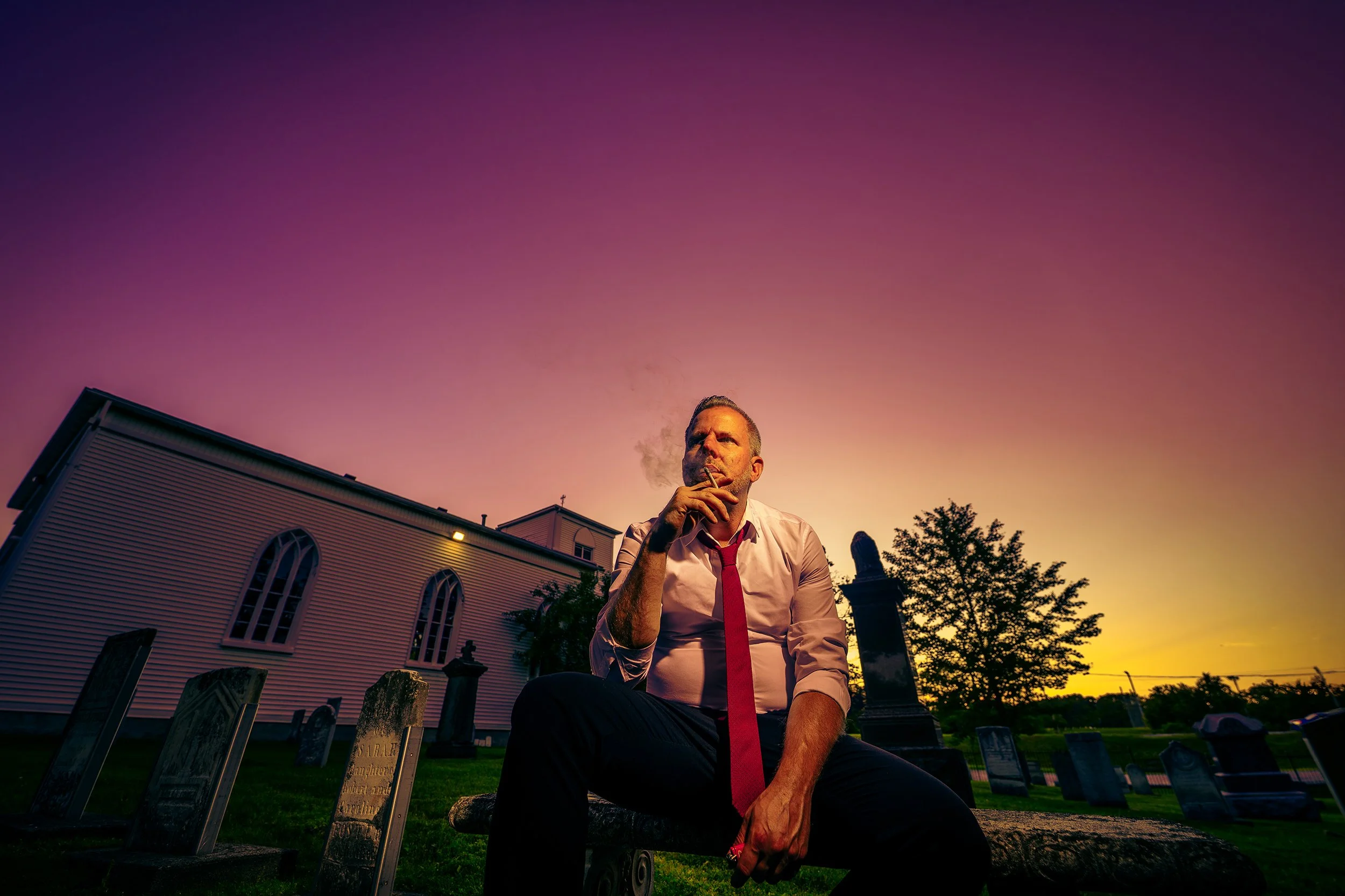 A middle-aged man dressed in a white shirt and red tie is sitting on a bench in a cemetery at sunset, smoking a cigarette. The church building and gravestones are visible behind him, with a colorful sky overhead.
