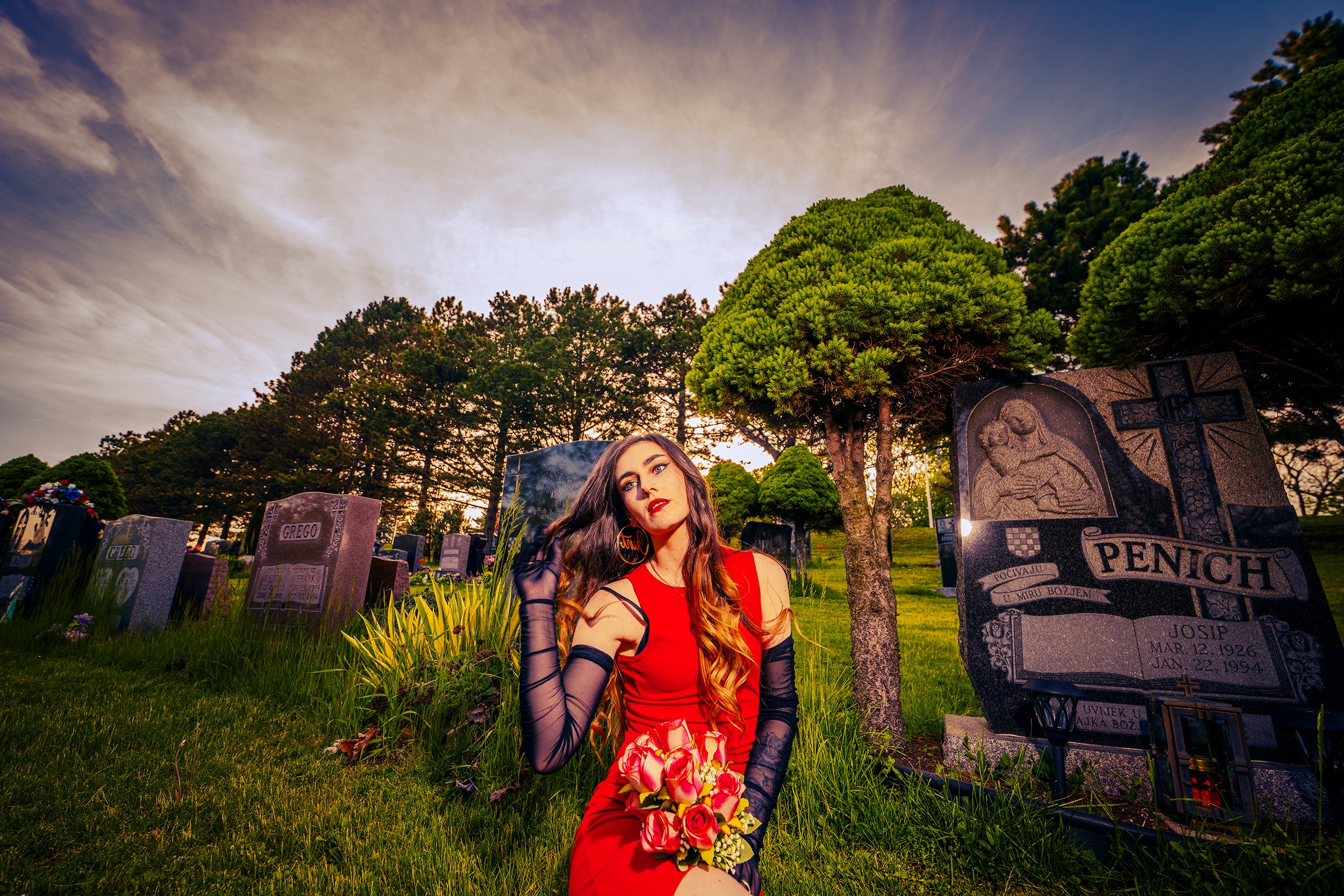 A woman in a red dress with black gloves and earrings sitting on the grass at a cemetery during sunset, holding a bouquet of pink and white roses, with tombstones and trees in the background.