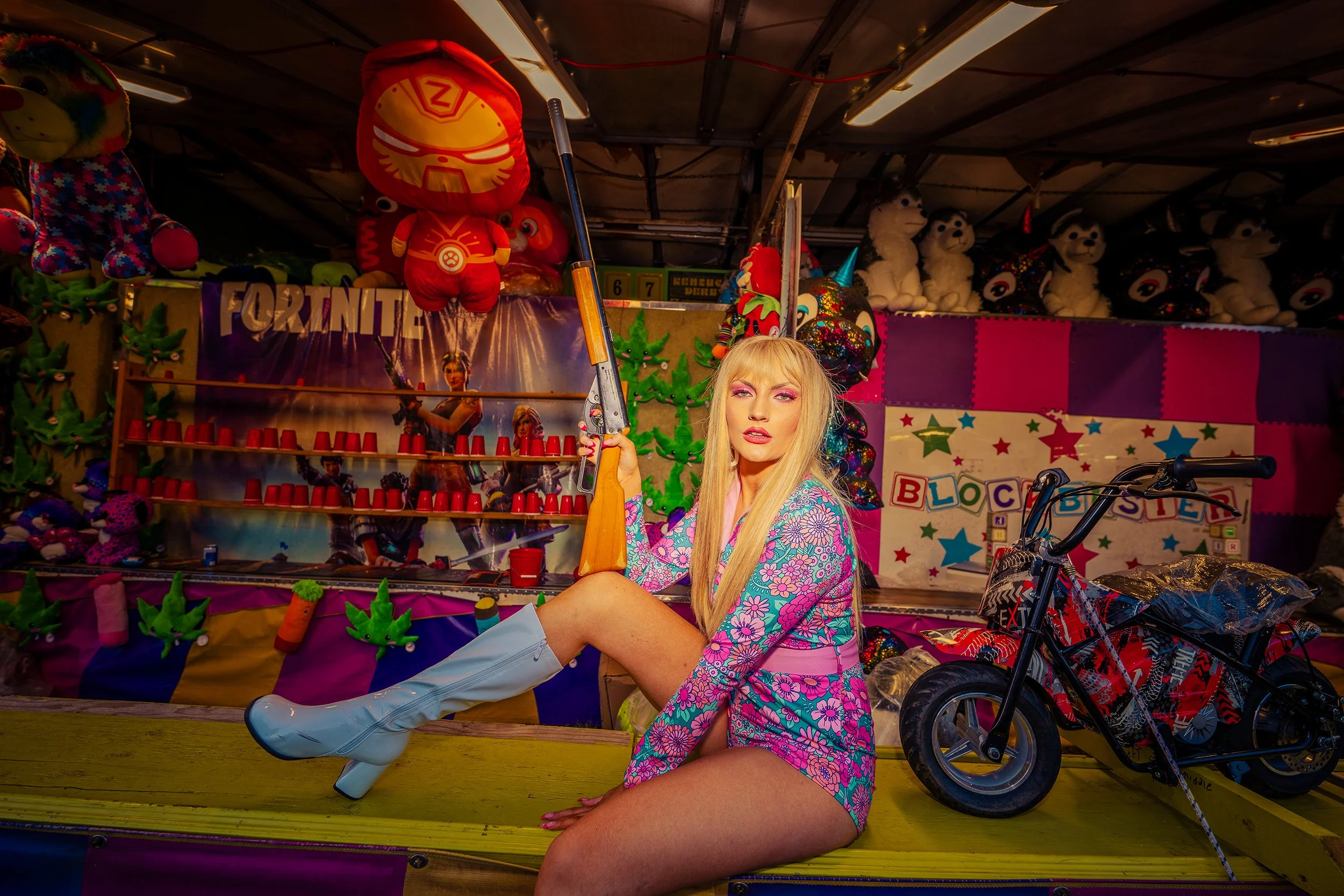 A woman with long blonde hair, wearing a colorful floral dress and silver knee-high boots, is sitting on a platform at a carnival game booth. She is holding a toy rifle, with a toy motorcycle beside her. The booth is decorated with plush toys, balloo