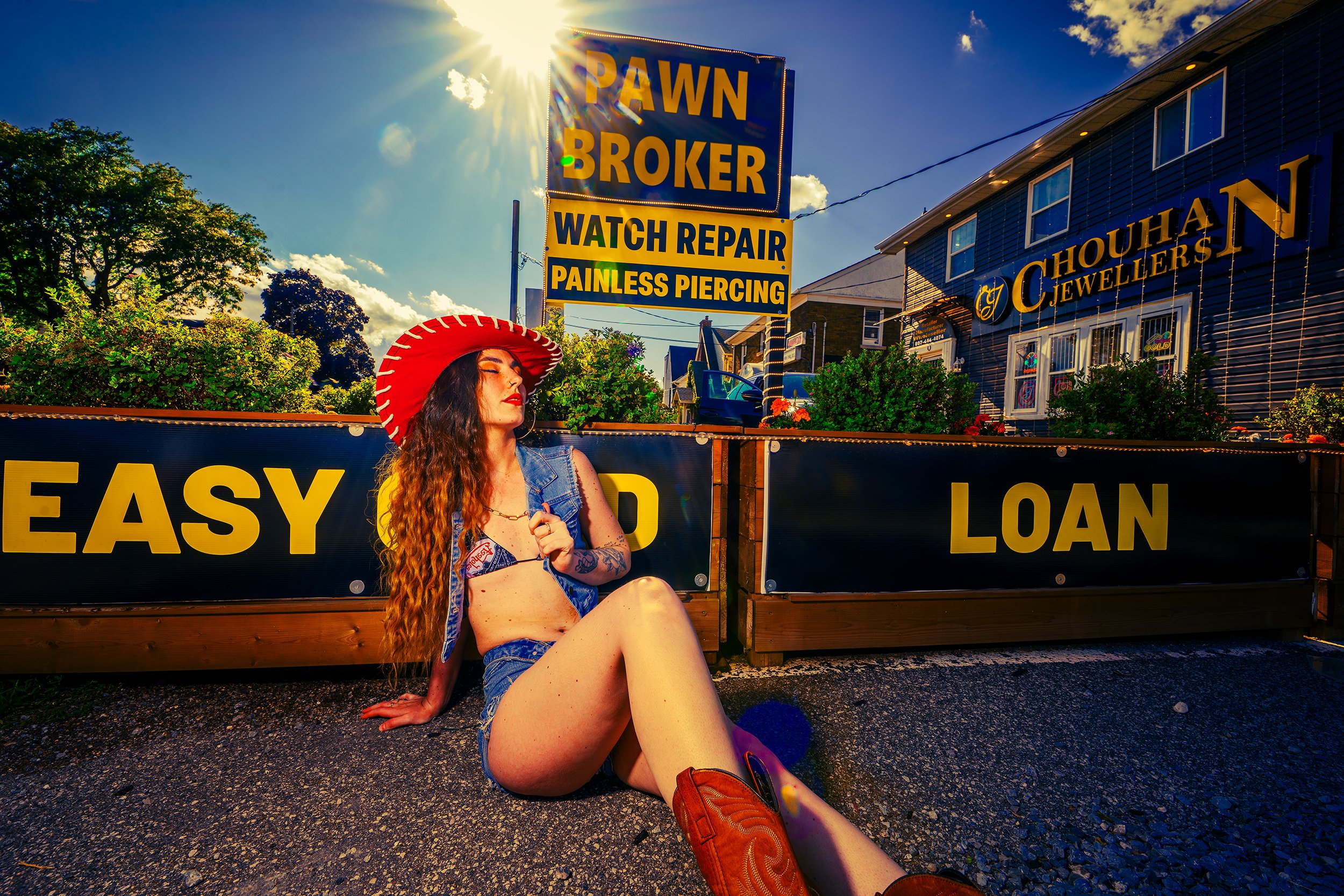A young woman with long red hair, tattoos, and wearing a denim vest and shorts, sitting on the ground in front of a wooden fence. She has a red wide-brimmed hat and red boots. Behind her, there are signs advertising pawn shop and jewelry services, an
