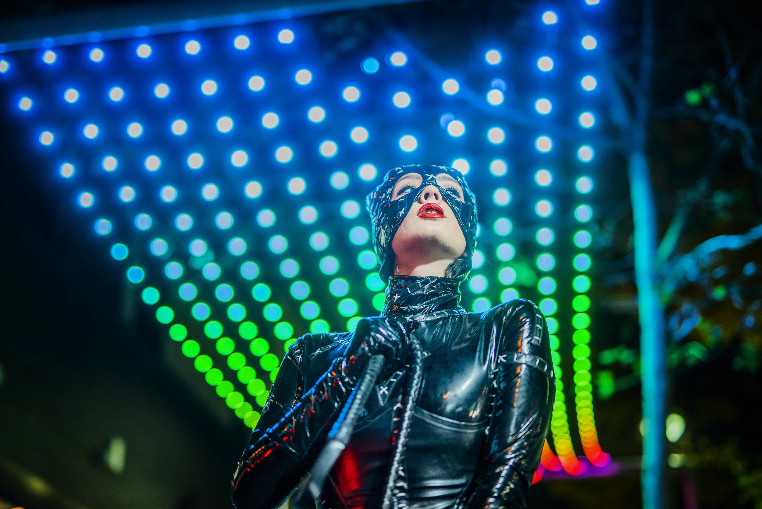 A person wearing a shiny black outfit with a matching face covering stands in front of a vibrant light display with blue and green circular lights forming a tunnel-like background at night.