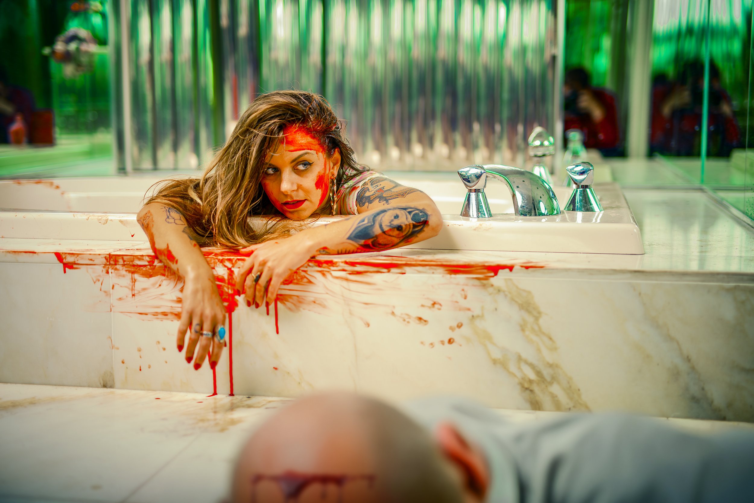 A woman with tattoos and smeared red makeup or paint on her face is lying in a bathtub with blood-like red streaks on the edge and in the water, with a distressed expression, in a room with green lighting and a reflective wall.