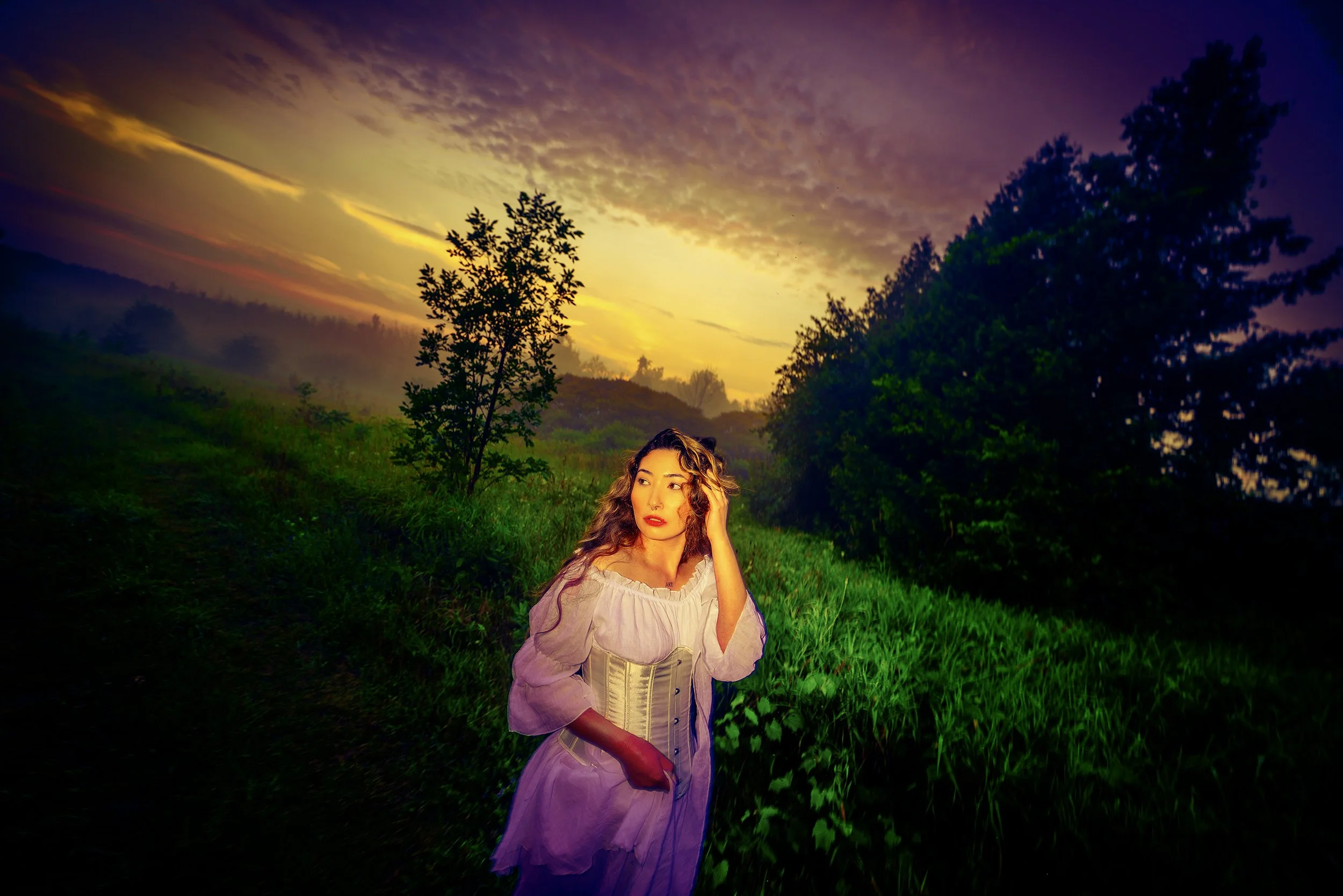 A woman with curly hair wearing a vintage-style dress in a field during sunset with a colorful, dramatic sky and trees in the background.