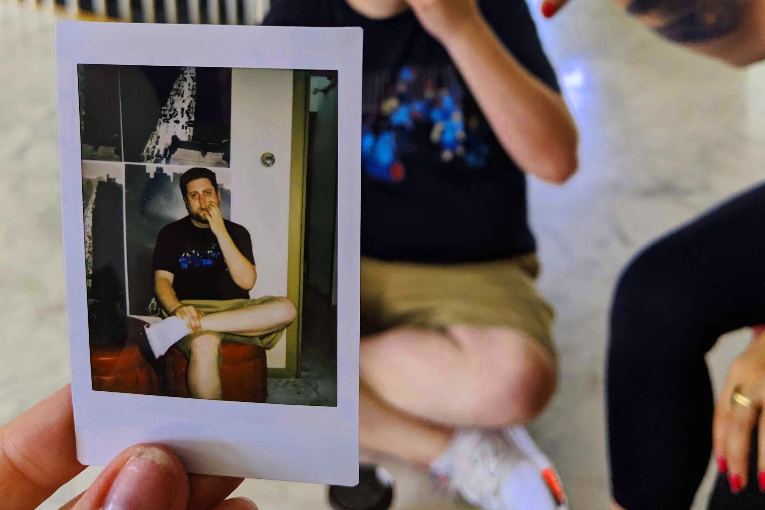 Photograph of a man sitting cross-legged with his hand on his chin, framed as a photo print being held in front of a person. The background shows an indoor space with a yellow wall and some first floor objects. The main figure appears to be young wit