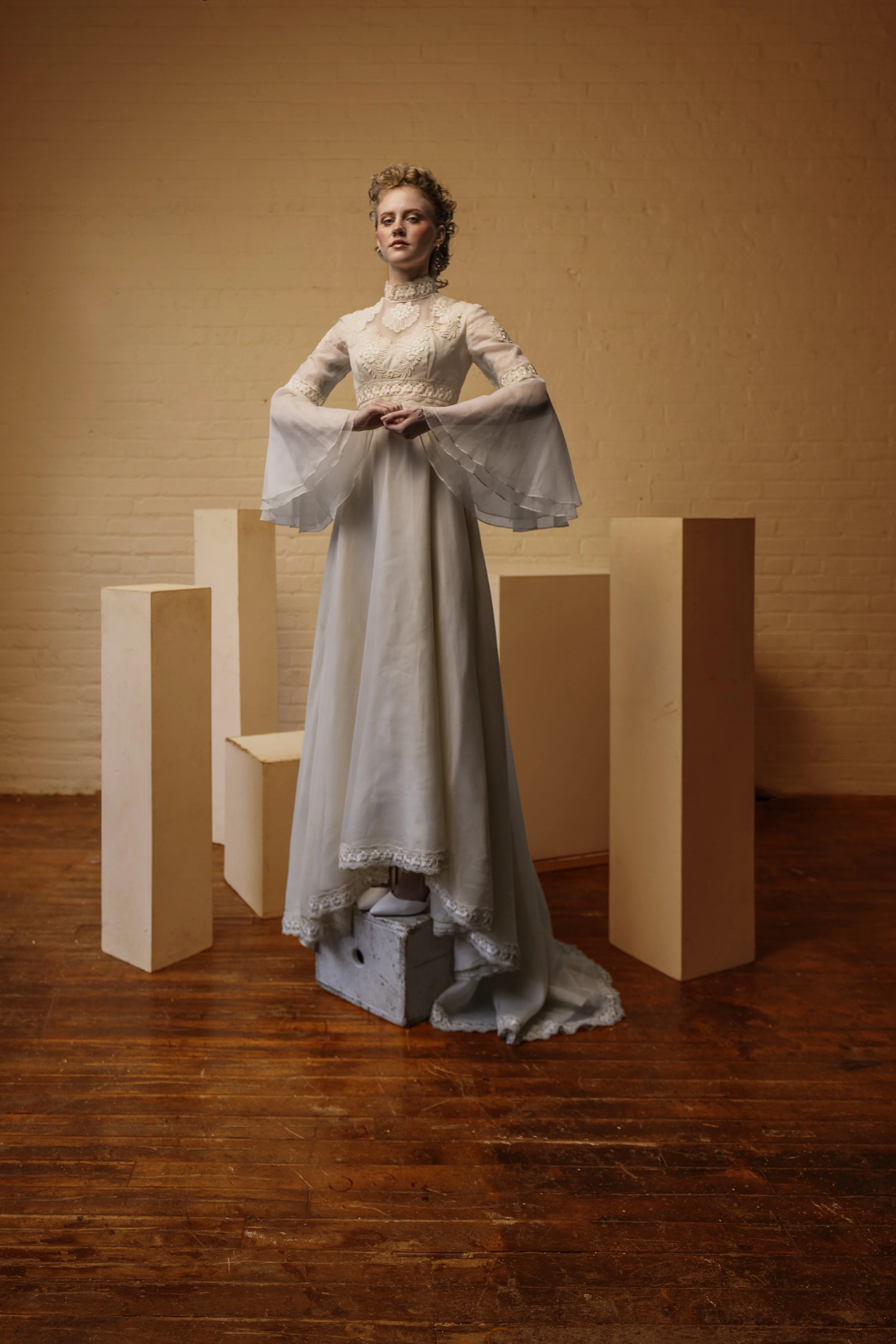 A woman dressed in a vintage white lace wedding gown standing on a small block in an art gallery, with light-colored blocks behind her against a beige brick wall.