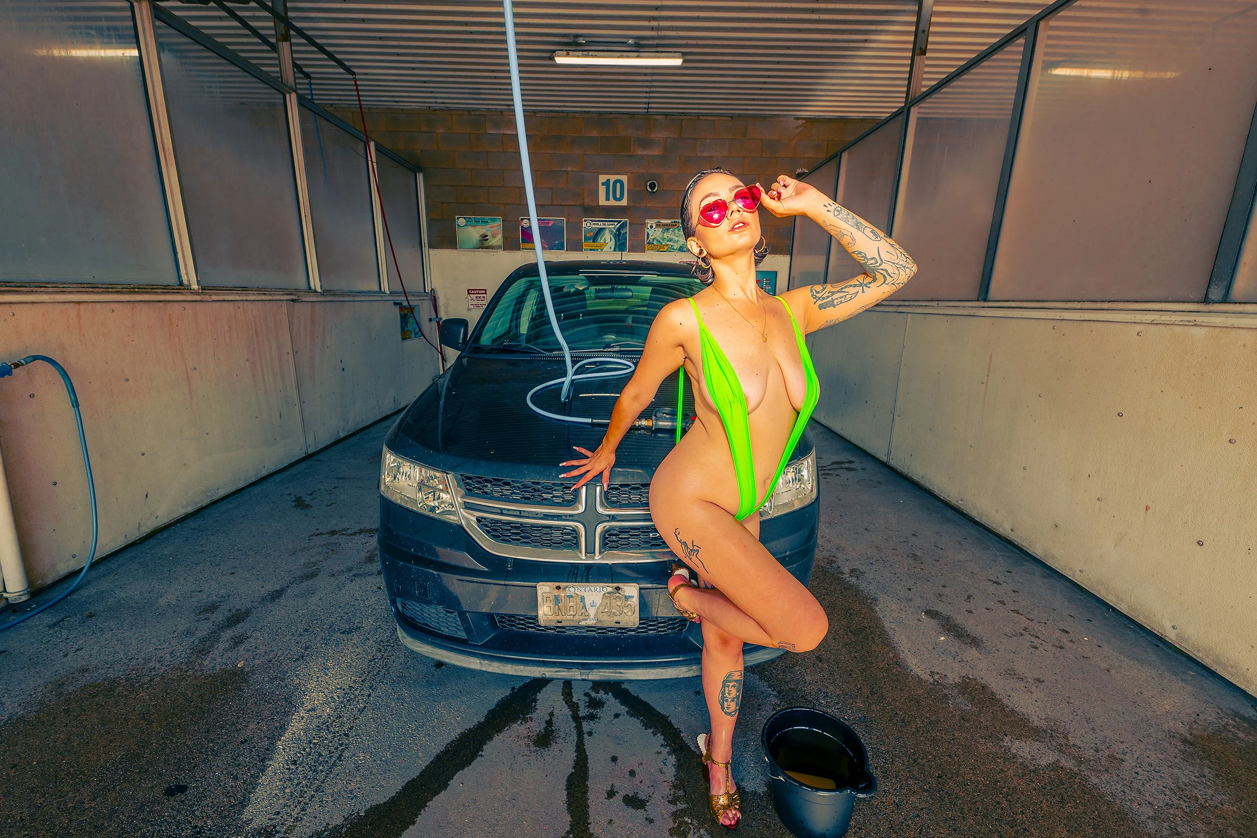 A woman in a green swimsuit and pink sunglasses posing in a car wash bay behind a black minivan. She has tattoos on her arms and legs, and is standing on one leg with her other knee bent, touching her sunglasses with one hand.