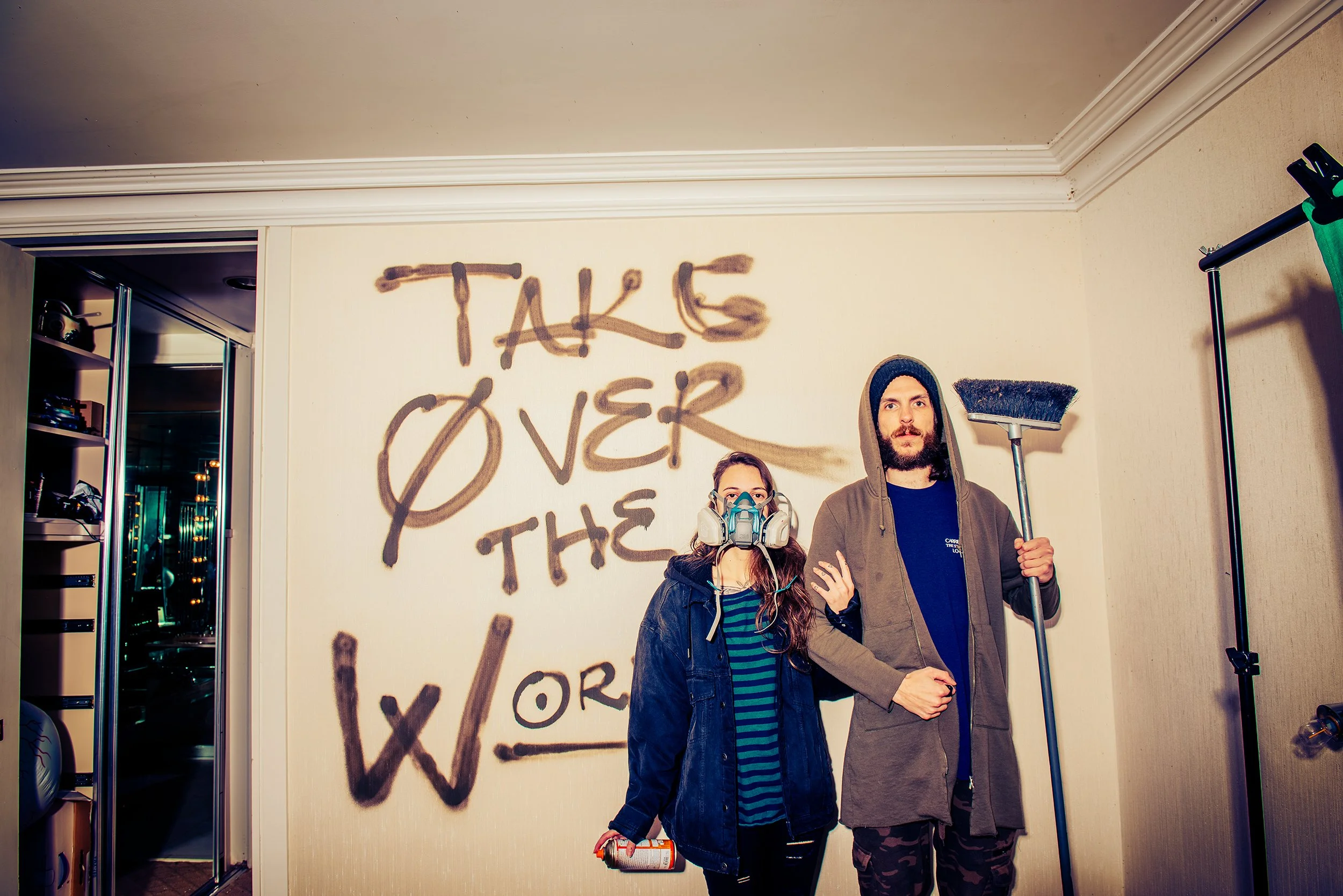 Two people standing in front of a wall with graffiti that says 'TAKE OVER THE' with the rest obscured. They are holding cleaning tools, and one person is wearing a mask.